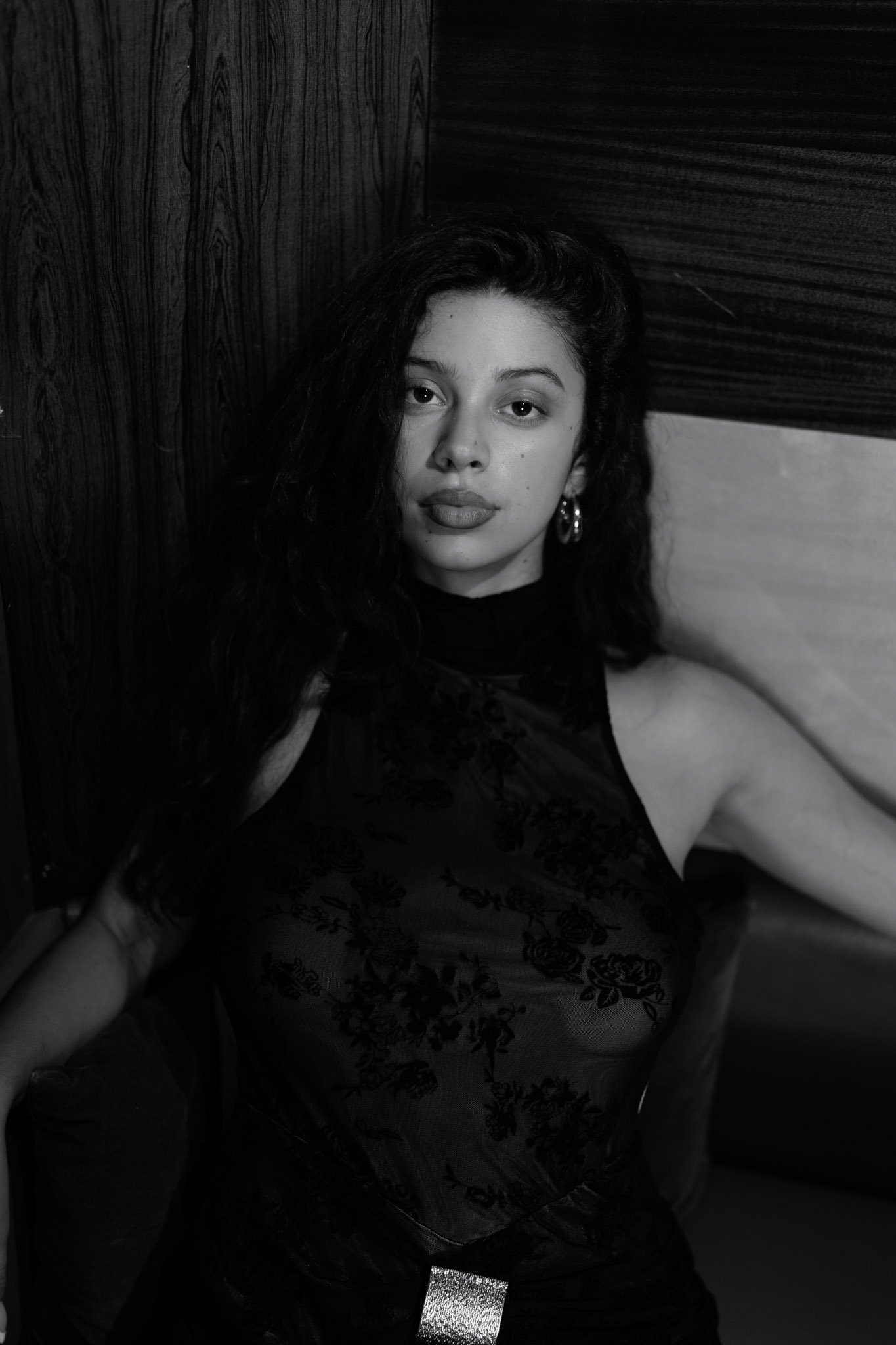 Black and white photo of a woman with dark curly hair, wearing earrings and a lace dark top, sitting against a wooden wall.