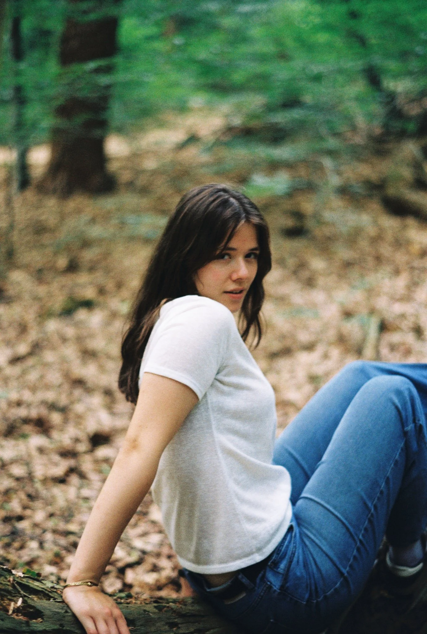 A young woman with long dark hair sitting on the ground in a wooded area, wearing a white t-shirt and blue jeans, looking towards the camera.