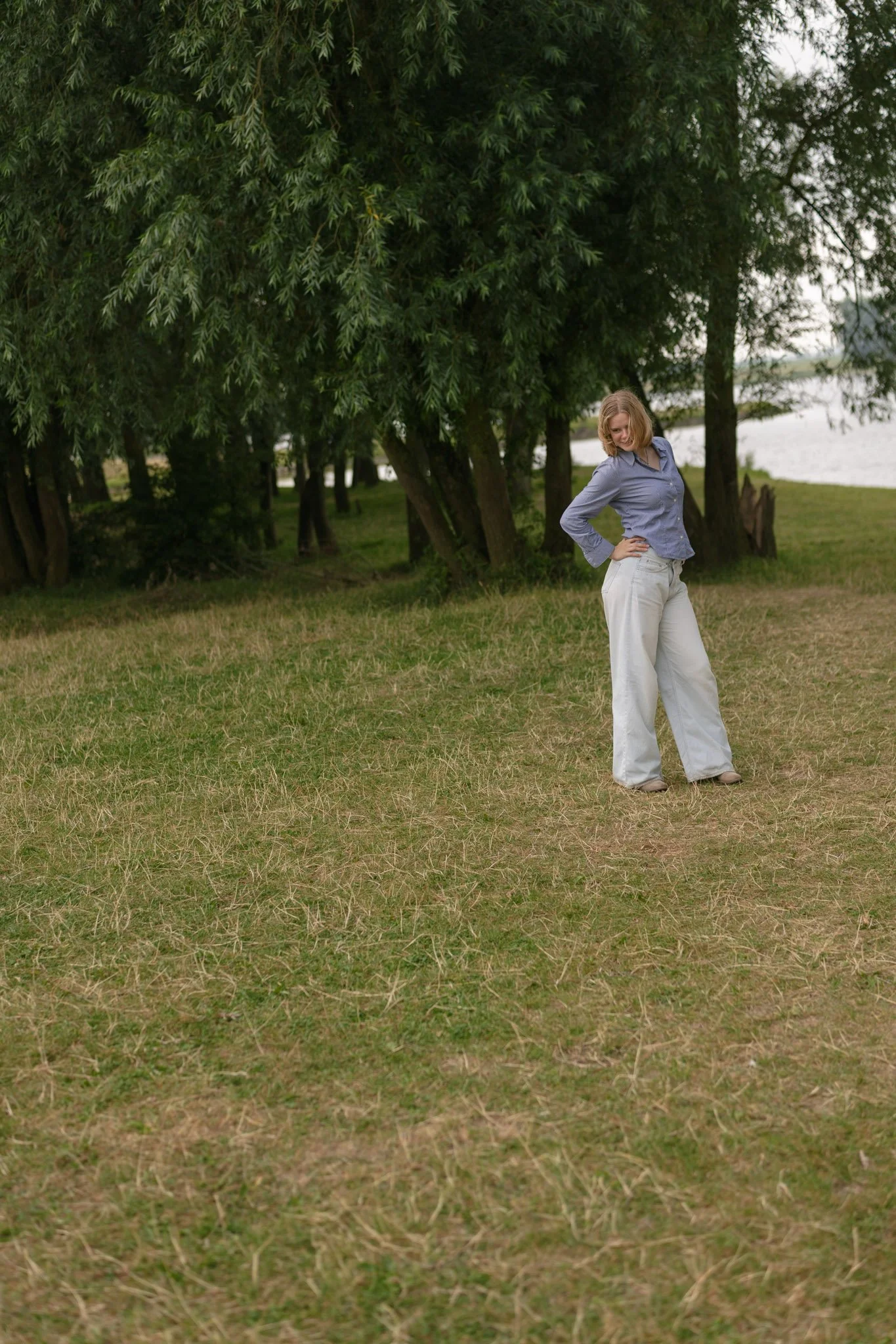 A woman with blonde hair wearing a blue button-up shirt and white pants is standing on a grassy area near trees and a body of water, looking down and smiling.