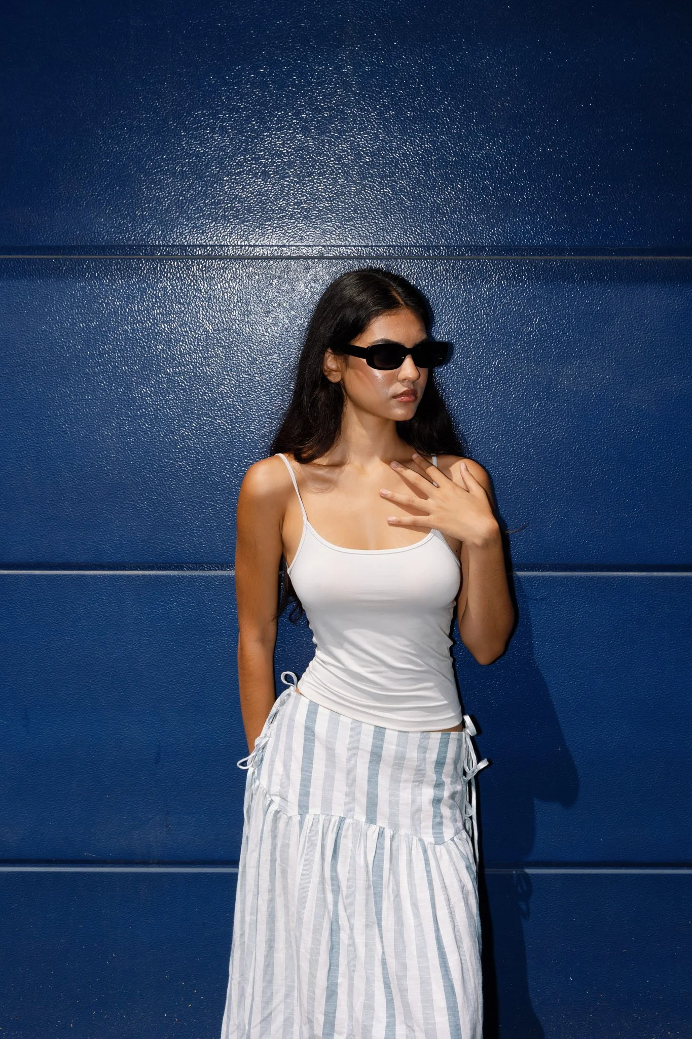 Young woman with long dark hair wearing sunglasses, a white tank top, and a striped skirt, standing against a blue wall.