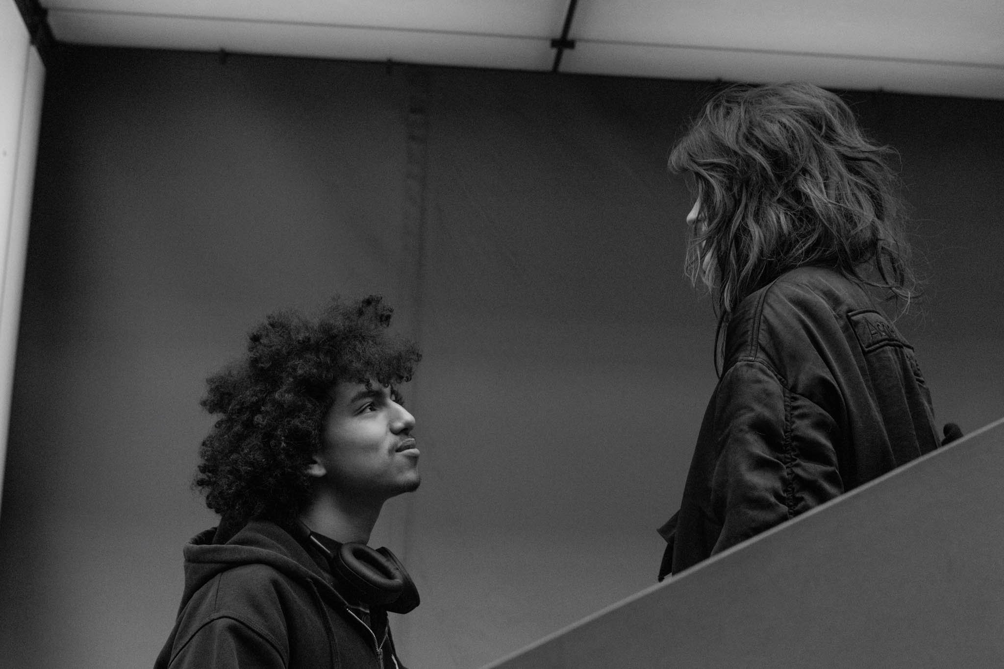 A black-and-white photo of a young man with curly hair looking at a woman with wavy hair. The woman is wearing a leather jacket and standing above the young man, who is also dressed in dark clothing. They are in an indoor setting, possibly a stairwell, with a plain wall and ceiling visible.