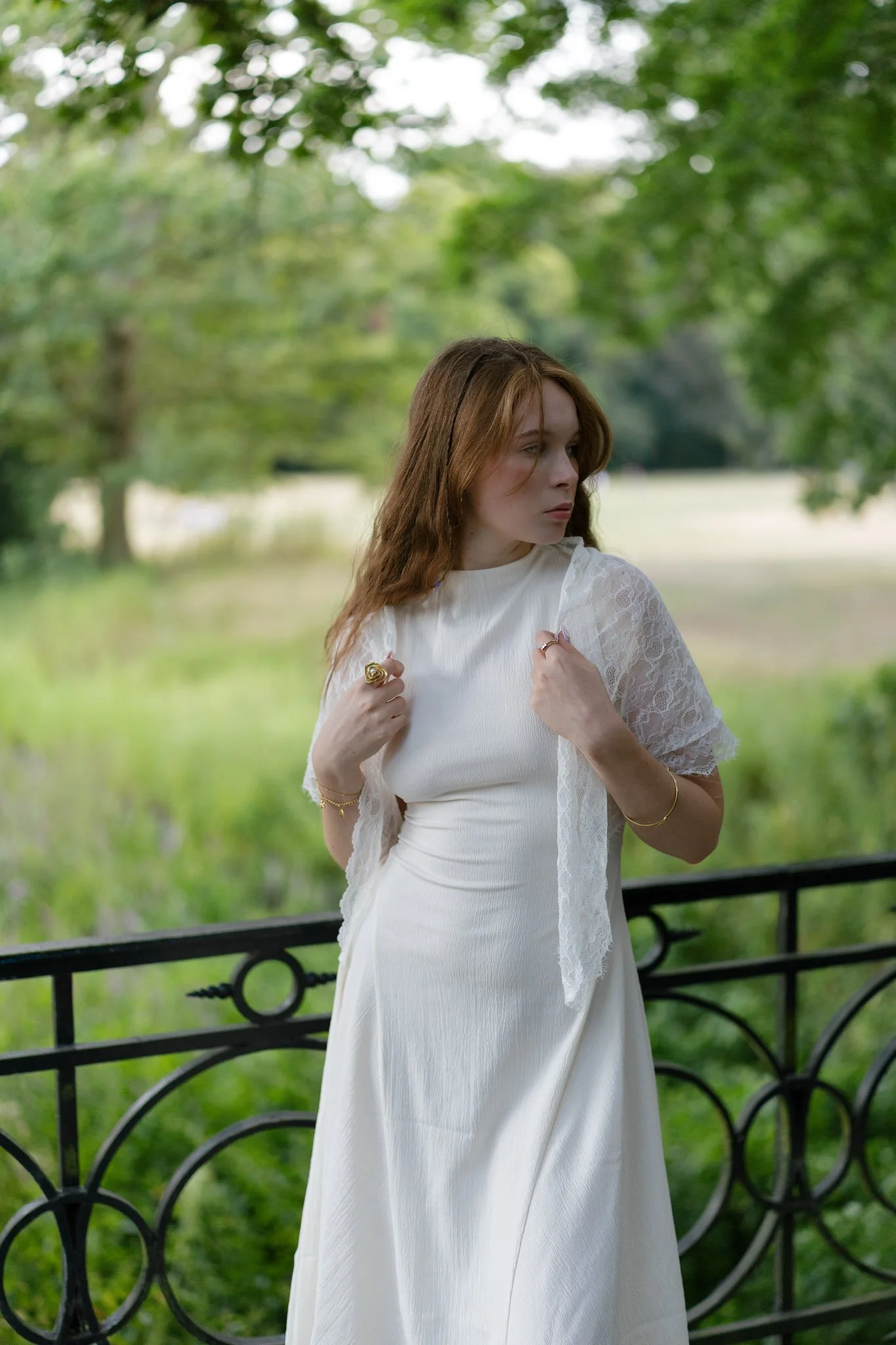 A young woman with auburn hair in a white dress with lace sleeves standing in a park with green trees and grass, looking to her right, holding her dress at the neckline with both hands.