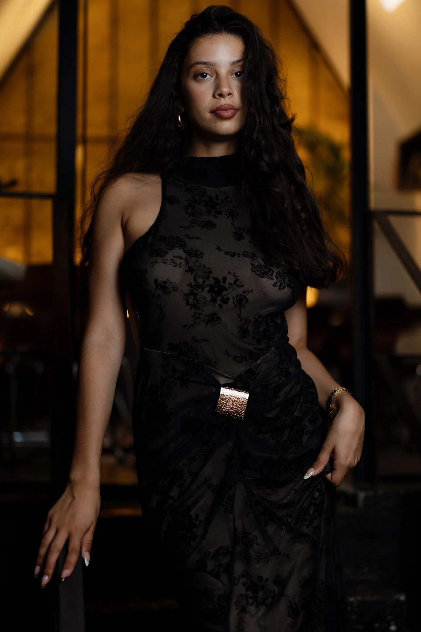 A woman with long, curly dark hair wearing a black, sheer, floral-patterned dress standing in a warmly lit indoor setting with wooden and black metal architectural elements in the background.