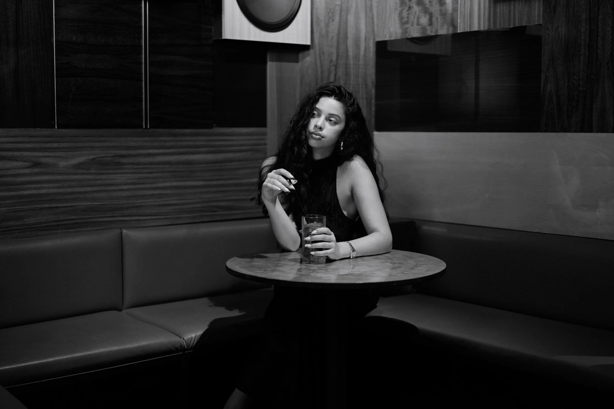 A young woman with long curly hair and earrings sits alone at a round table in a dark, modern restaurant. She is holding a glass of dark beverage and appears to be looking thoughtfully to the side.