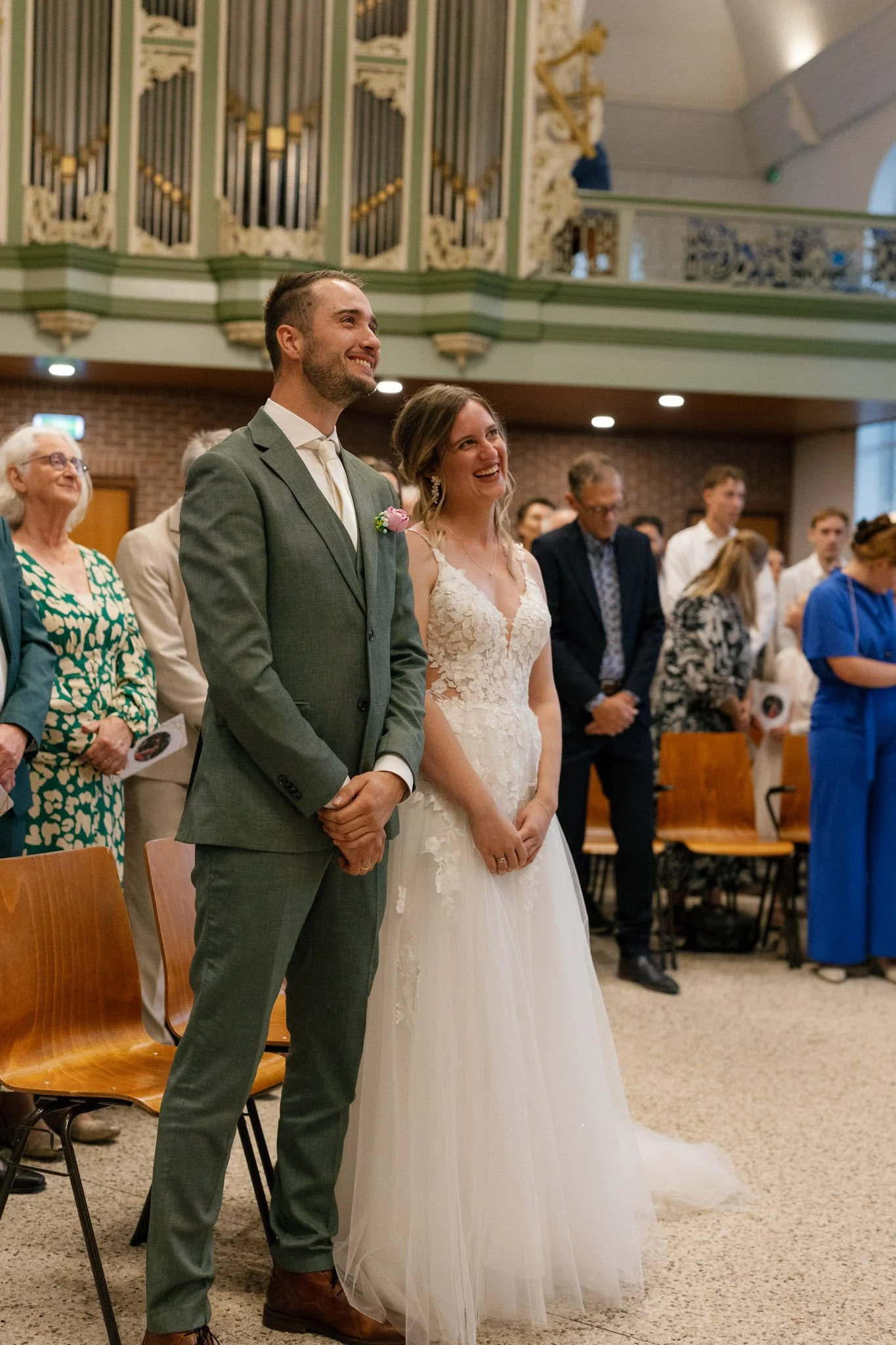 A bride and groom standing together at their wedding ceremony, smiling and looking forward, with guests in the background inside a decorated venue.