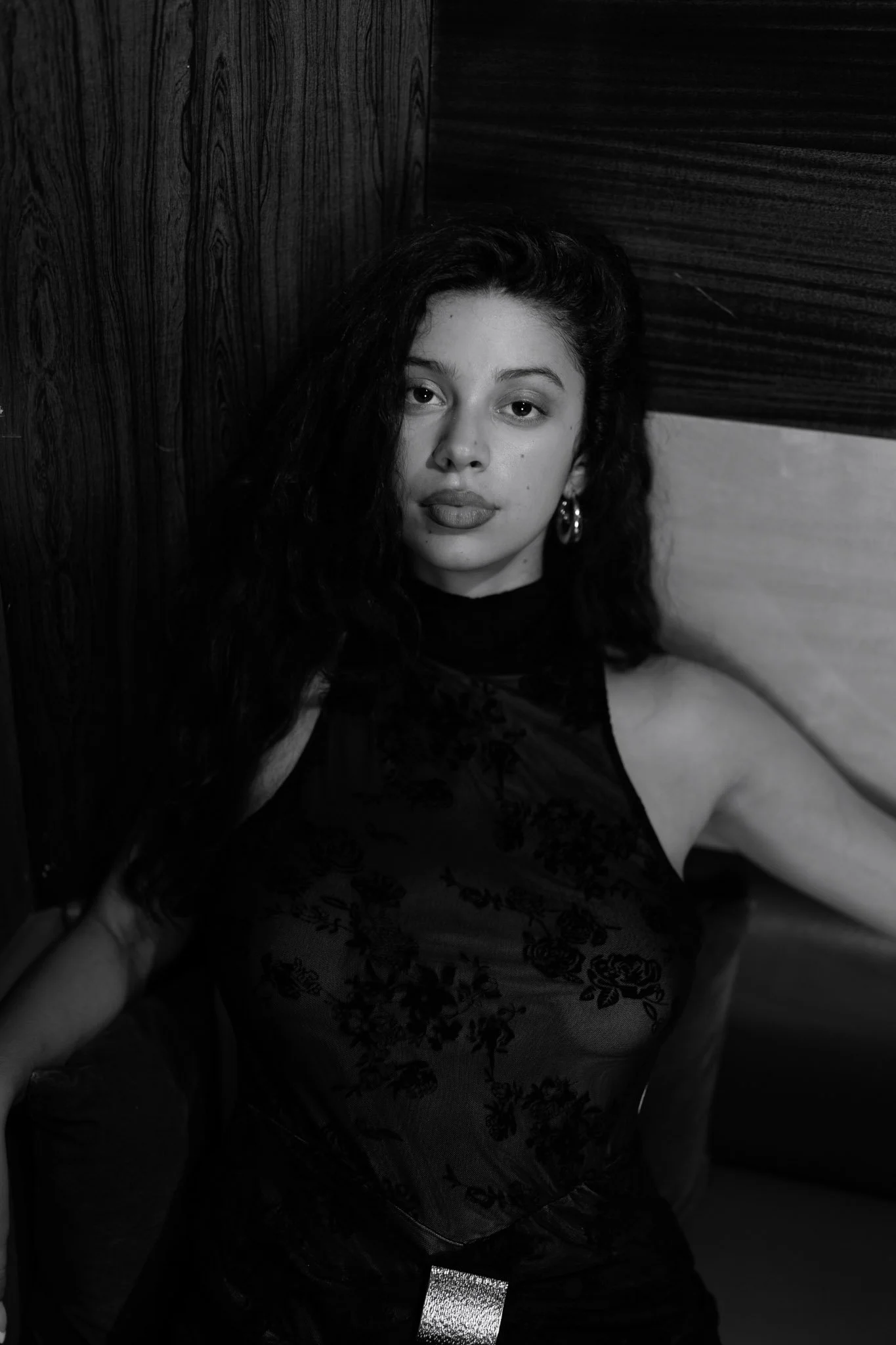 A woman with dark wavy hair and hoop earrings taking a selfie in a black and white photo, wearing a dark dress with floral embroidery.