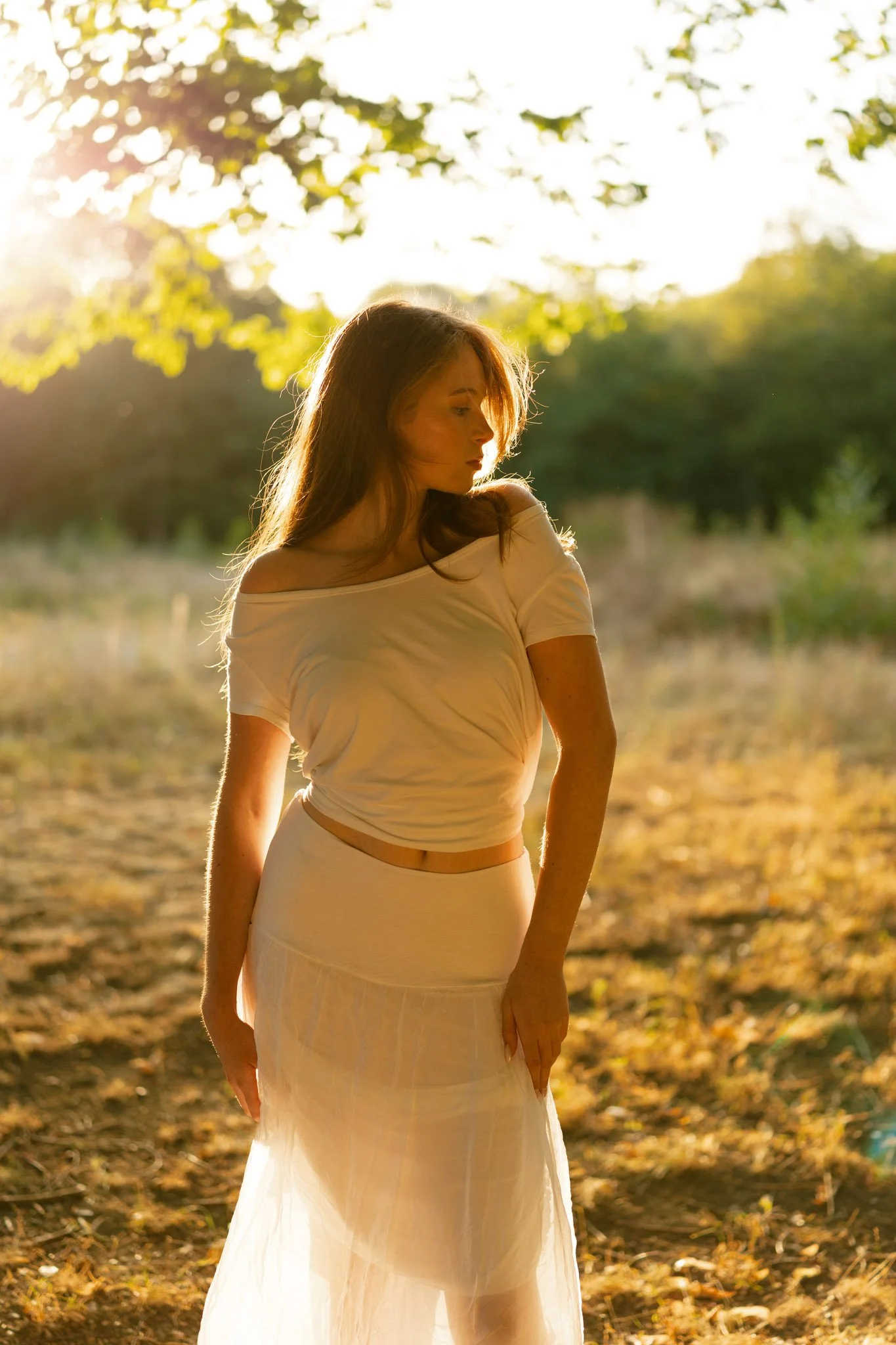 A young woman standing outdoors in a field during sunset, wearing a cream-colored off-shoulder top and a sheer white skirt, with sunlight filtering through the trees behind her.
