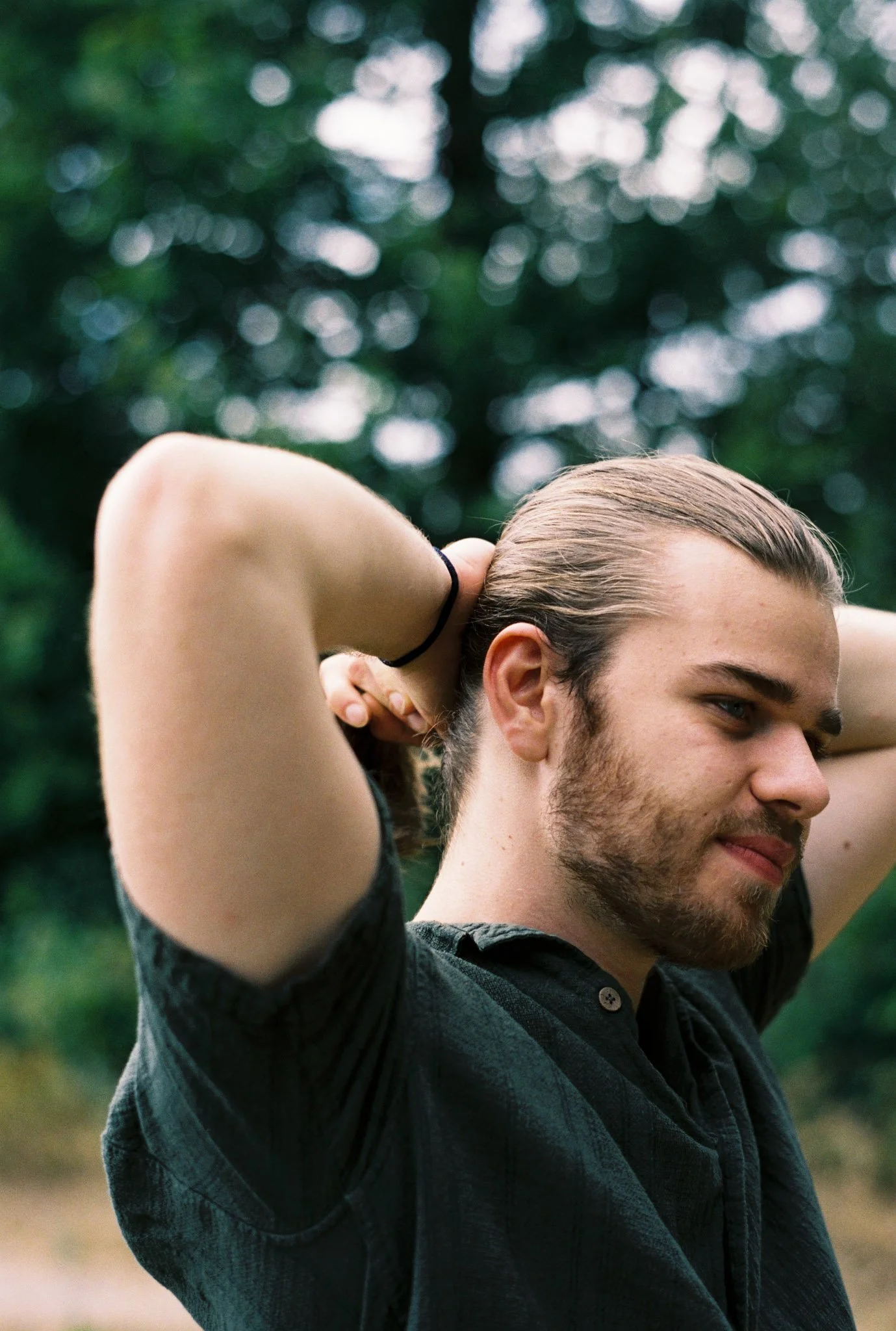 A young man with long hair and a beard is outdoors, adjusting his hair with both hands on the back of his head, in front of a blurred green and white background.