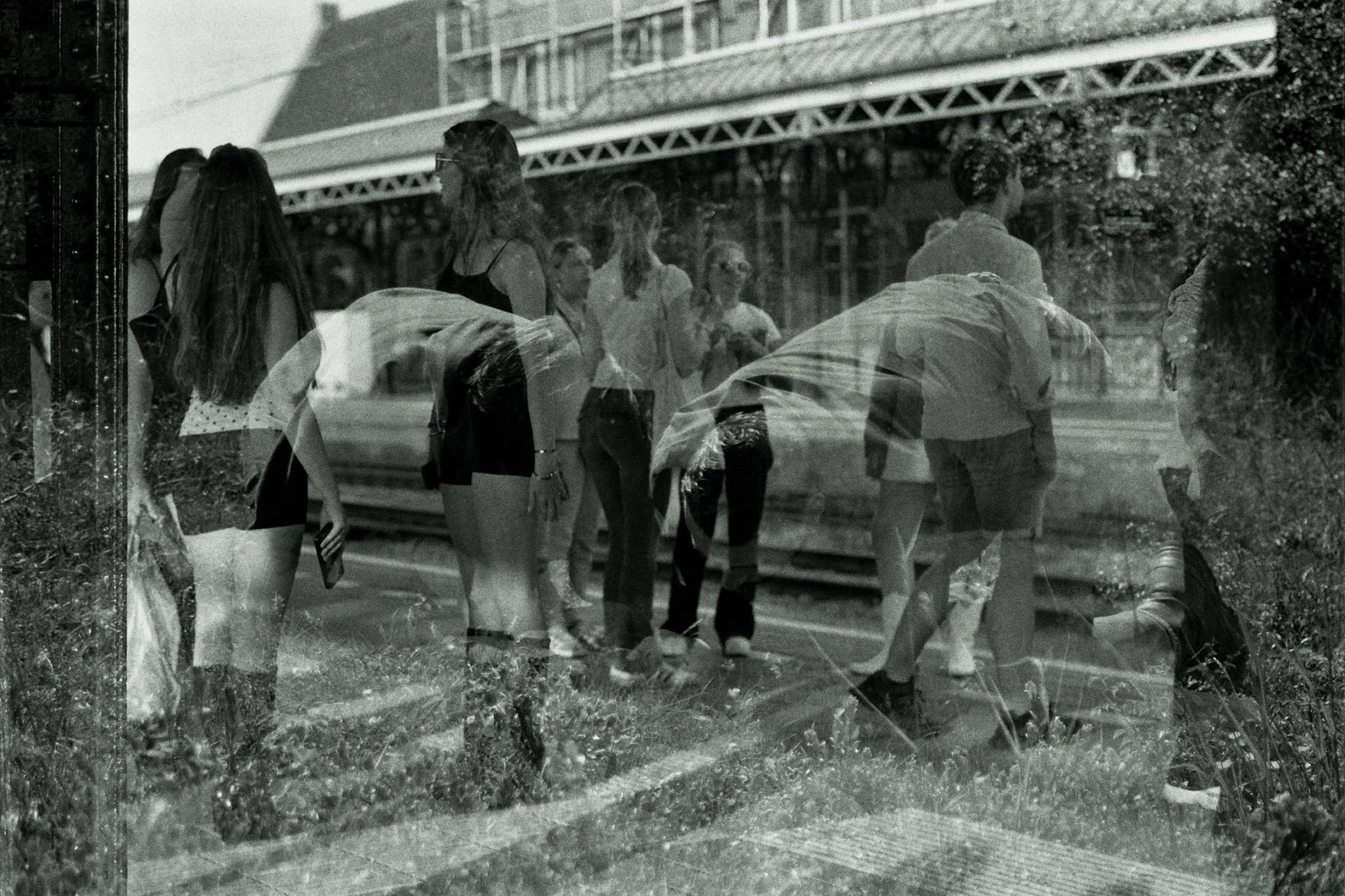A reflection of several people, mostly young women, standing and walking near a railroad track with an overpass or bridge structure in the background. The image appears to be taken through a glass window, giving it a layered, slightly blurry effect.