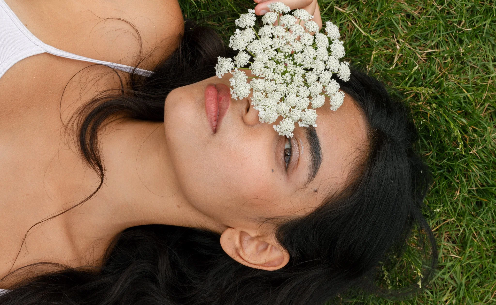 A woman with long dark hair lying on green grass, holding white flowers in front of her eyes.