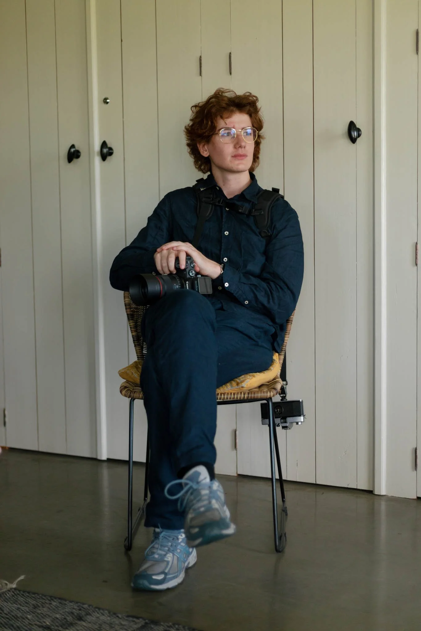 A person with curly red hair, glasses, and a serious expression sitting in a room with wooden paneled walls, holding a camera. They are wearing a dark shirt, jeans, and sneakers, with a backpack on their shoulders.