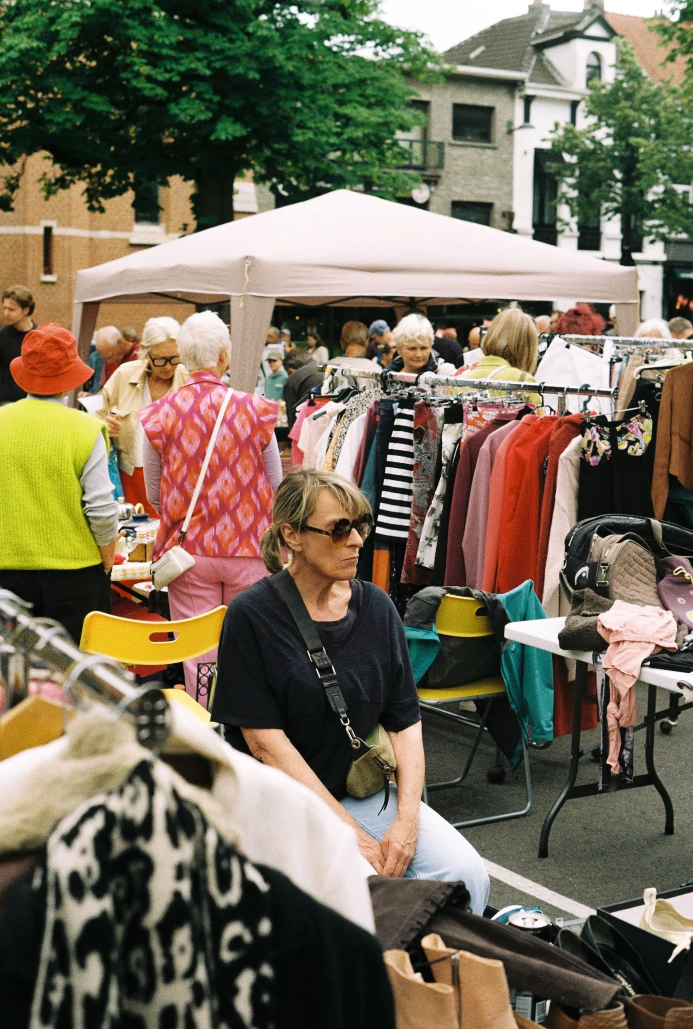 People browsing at an outdoor flea market with clothing racks, tables, and a white canopy, surrounded by trees and residential buildings.