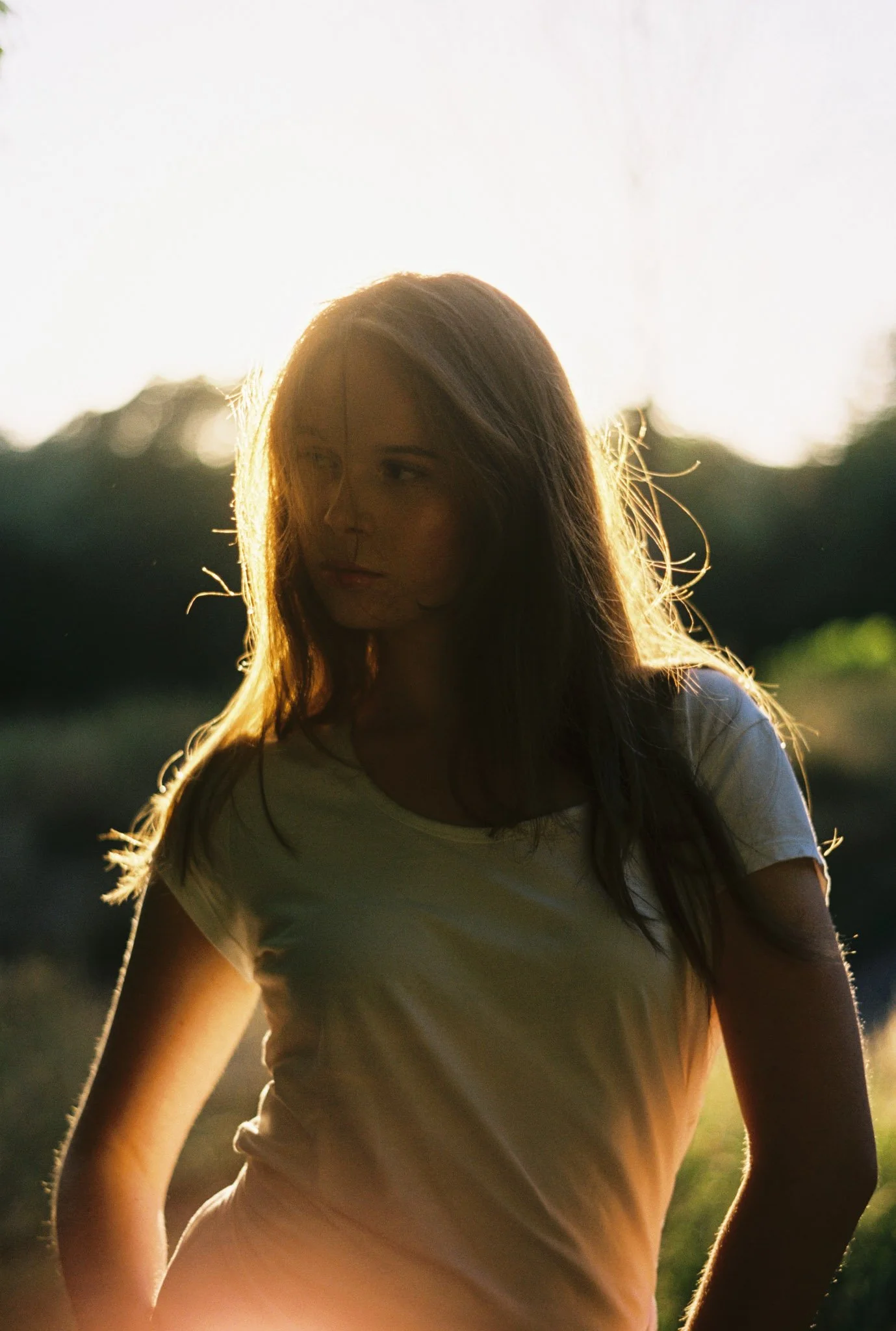 A woman with long hair wearing a white t-shirt outdoors during sunset.