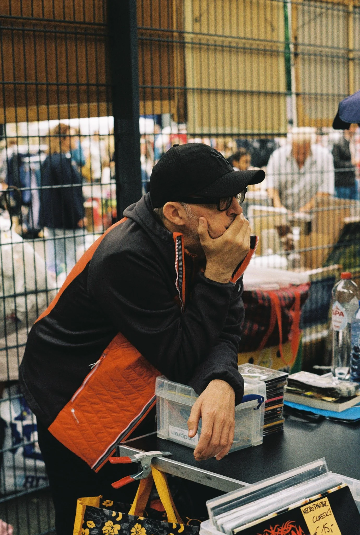 A man wearing glasses, a black cap, and a black and orange jacket at a table with vinyl records at a store or market, with other people and store shelves in the background.