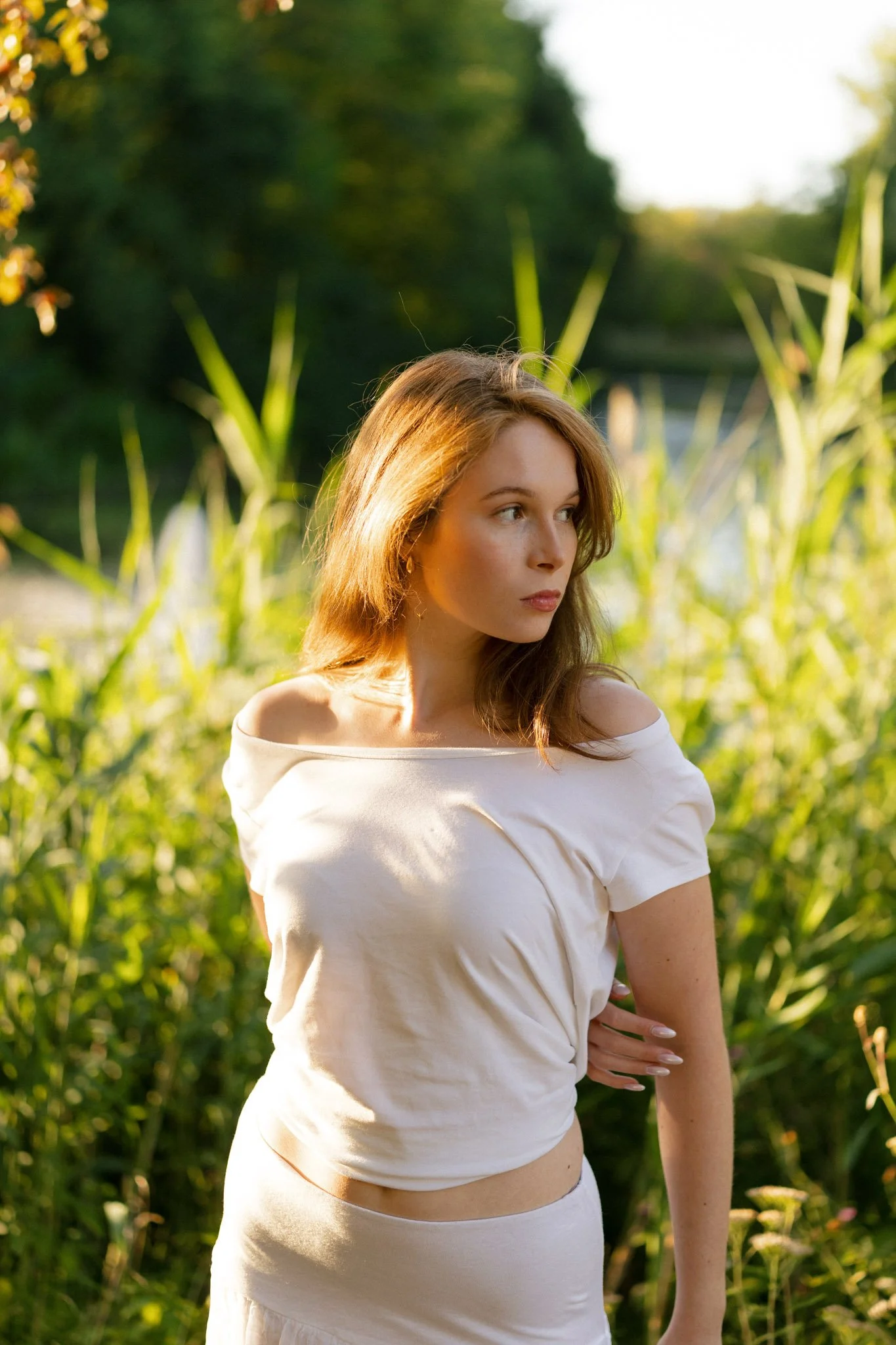A woman with long, light brown hair standing outdoors in greenery, wearing a white off-shoulder top and skirt, with sunlight illuminating part of her face and hair, and looking to her right.