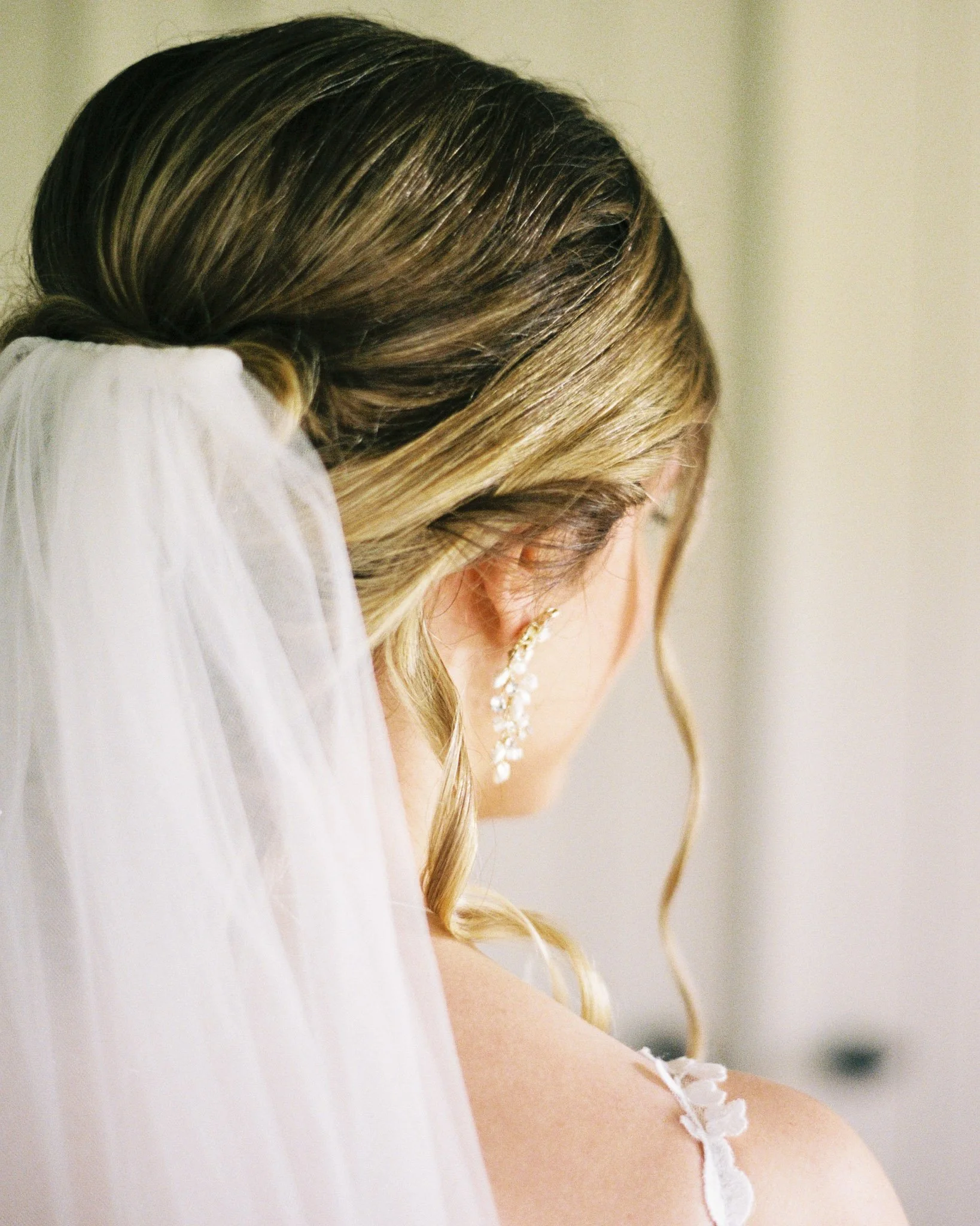 Close-up of a woman with blonde hair in French braid, wearing a white lace strap dress and dangling earrings, with a blurred neutral background.