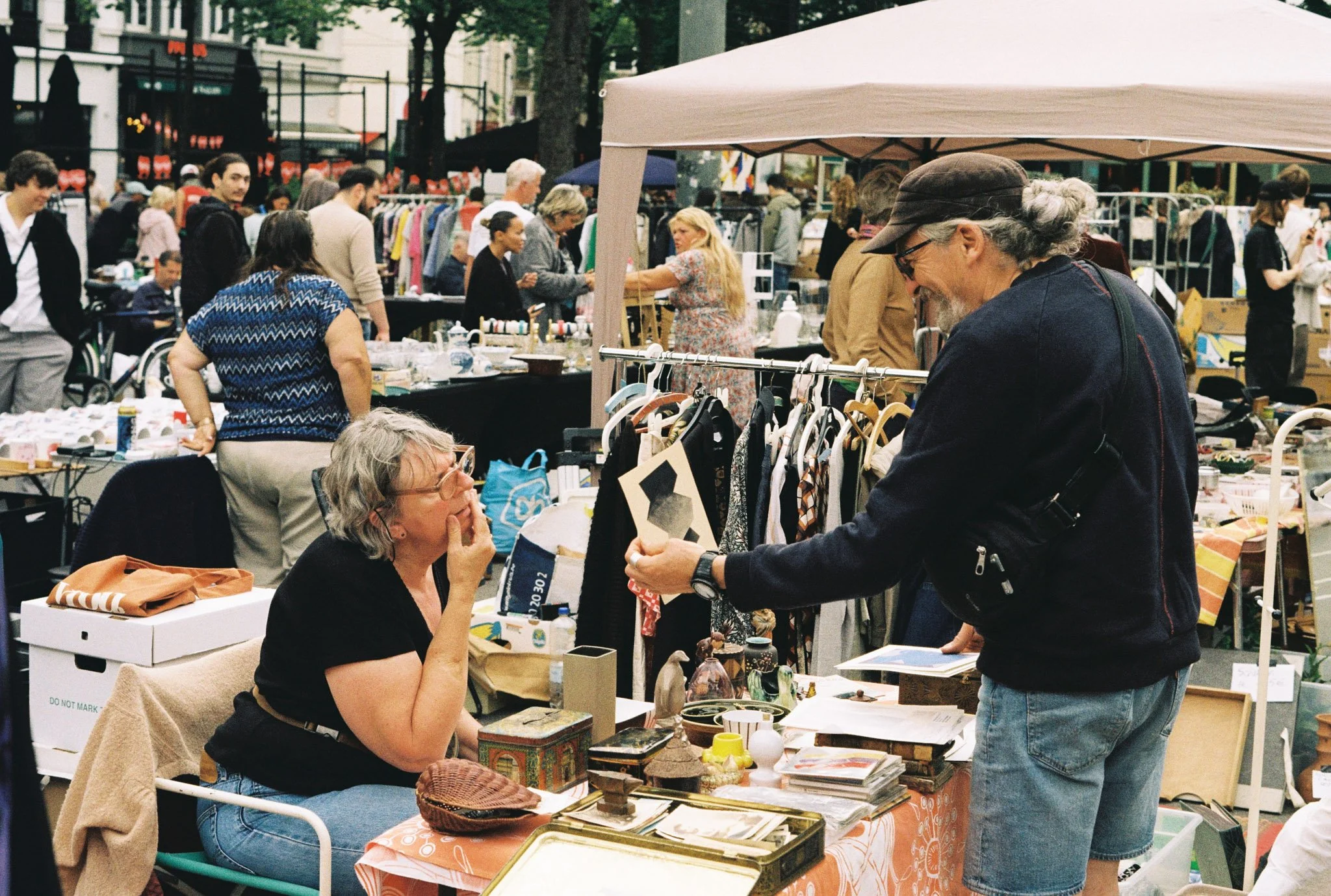 People shopping at an outdoor flea market, browsing various items displayed on tables and racks under tents.