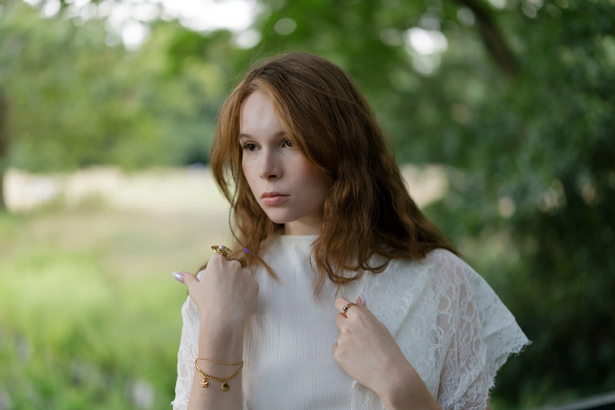 A young woman with wavy red hair standing outdoors in front of green trees, wearing a white lace dress, holding her hands near her chest, with rings and bracelets, looking off to the side with a calm expression.