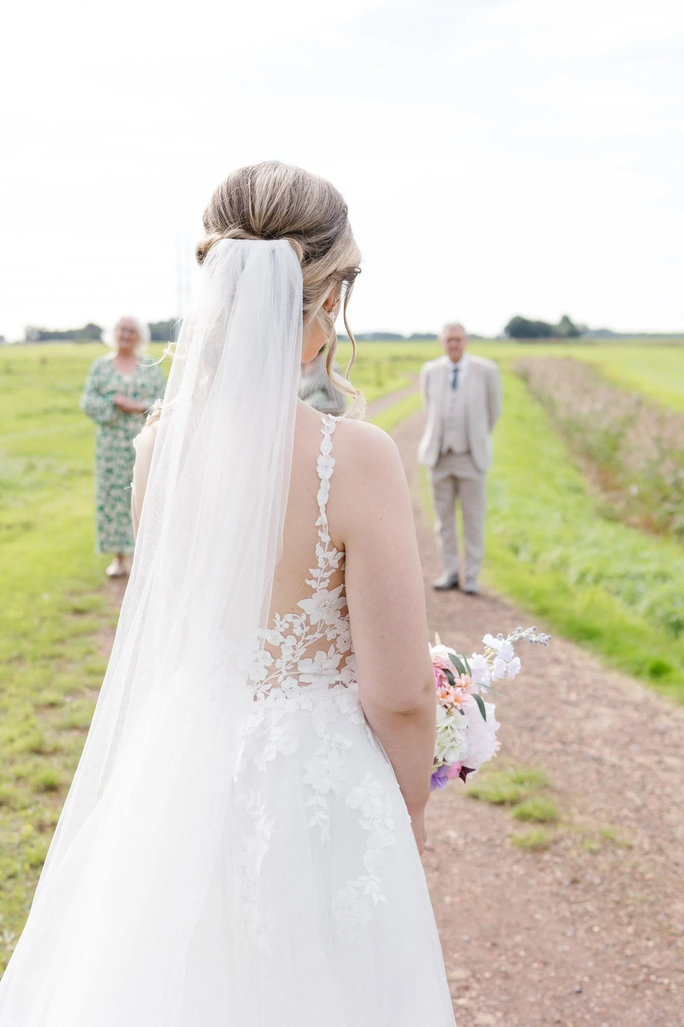 A bride in a white wedding dress with lace details holding a bouquet, standing outdoors on a dirt path, with family members in the background, during daytime.