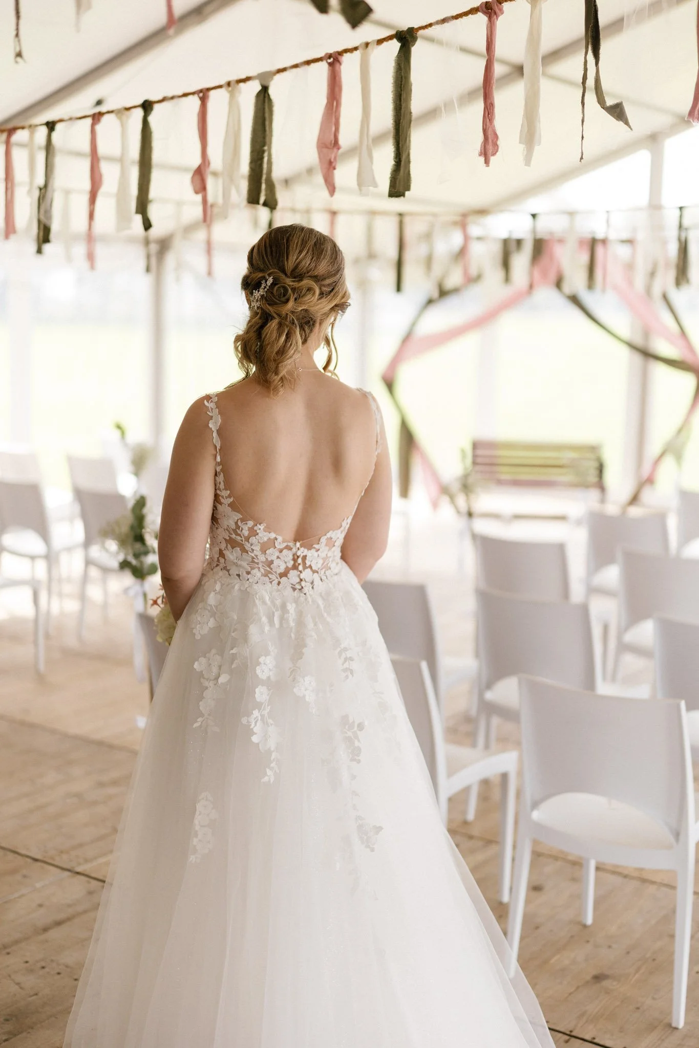 A bride in a wedding dress with floral lace details stands in an indoor wedding venue decorated with pink, white, and dark green ribbons hanging from a string, white chairs, and a wooden bench in the background.