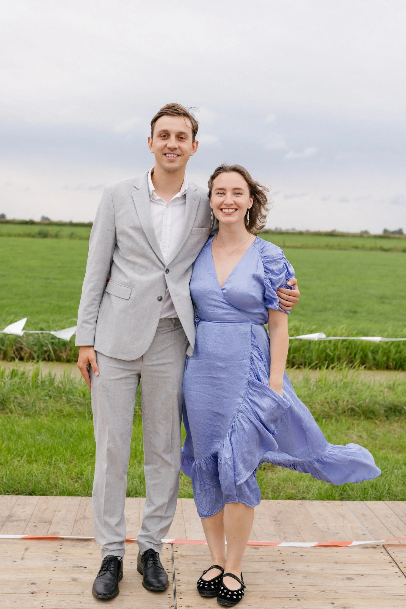 A man and woman smiling outdoors on a cloudy day, standing on a wooden platform with grassy fields in the background. The man wears a light gray suit, white shirt, and black shoes. The woman wears a purple dress and black shoes with white polka dots.