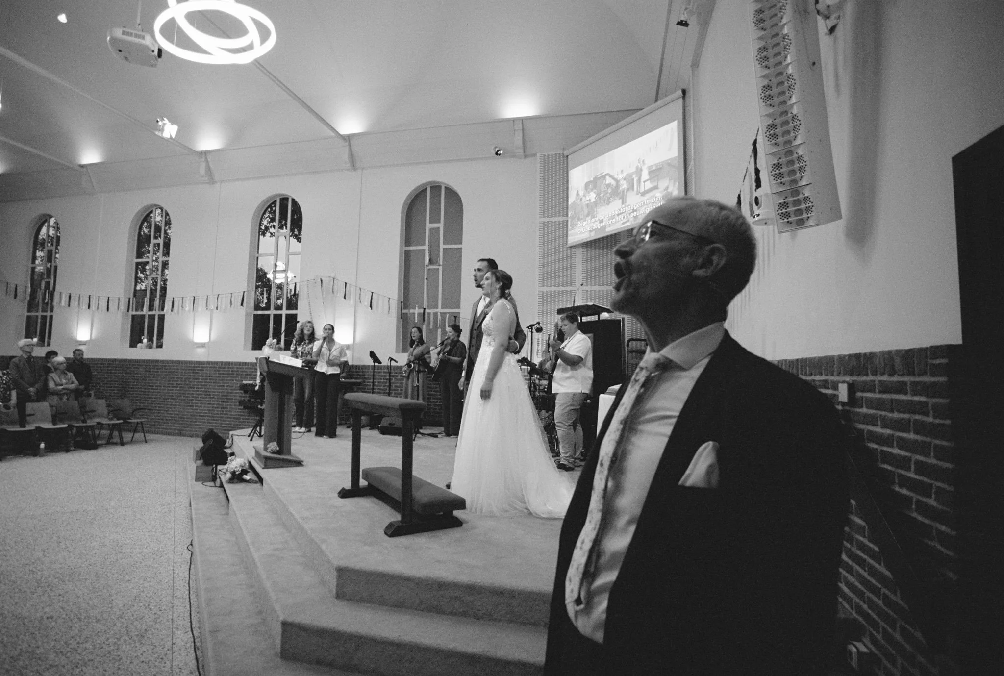 A black and white photo of a wedding ceremony inside a church. The bride and groom are standing together at the altar, with a singer or speaker and a band or choir nearby. Several guests are seated, and large windows with arches are visible in the background.