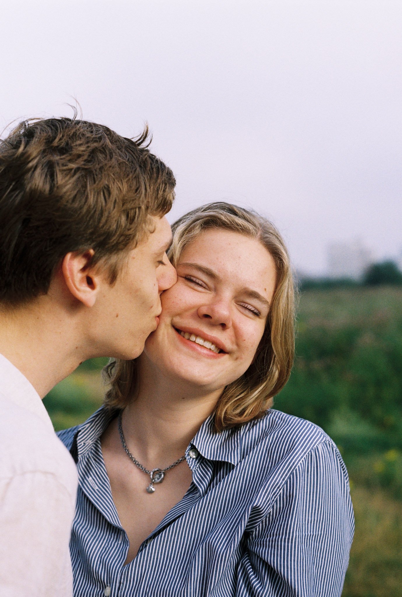 A young couple, a man and a woman, sharing a kiss outdoors in a field, with the woman smiling.