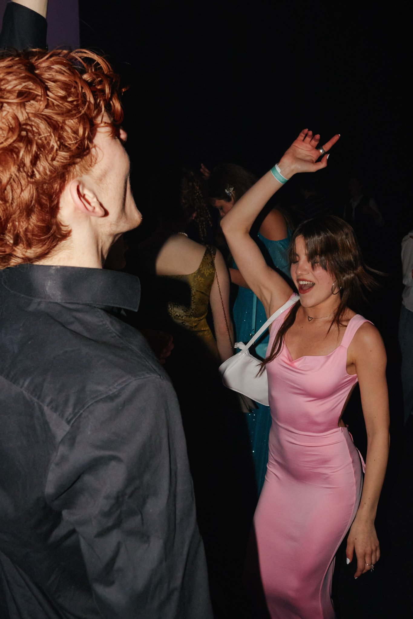 A woman with long brown hair wearing a pink satin dress dancing with her eyes closed, and a man with reddish curly hair and a black shirt dancing with friends in a dark setting.
