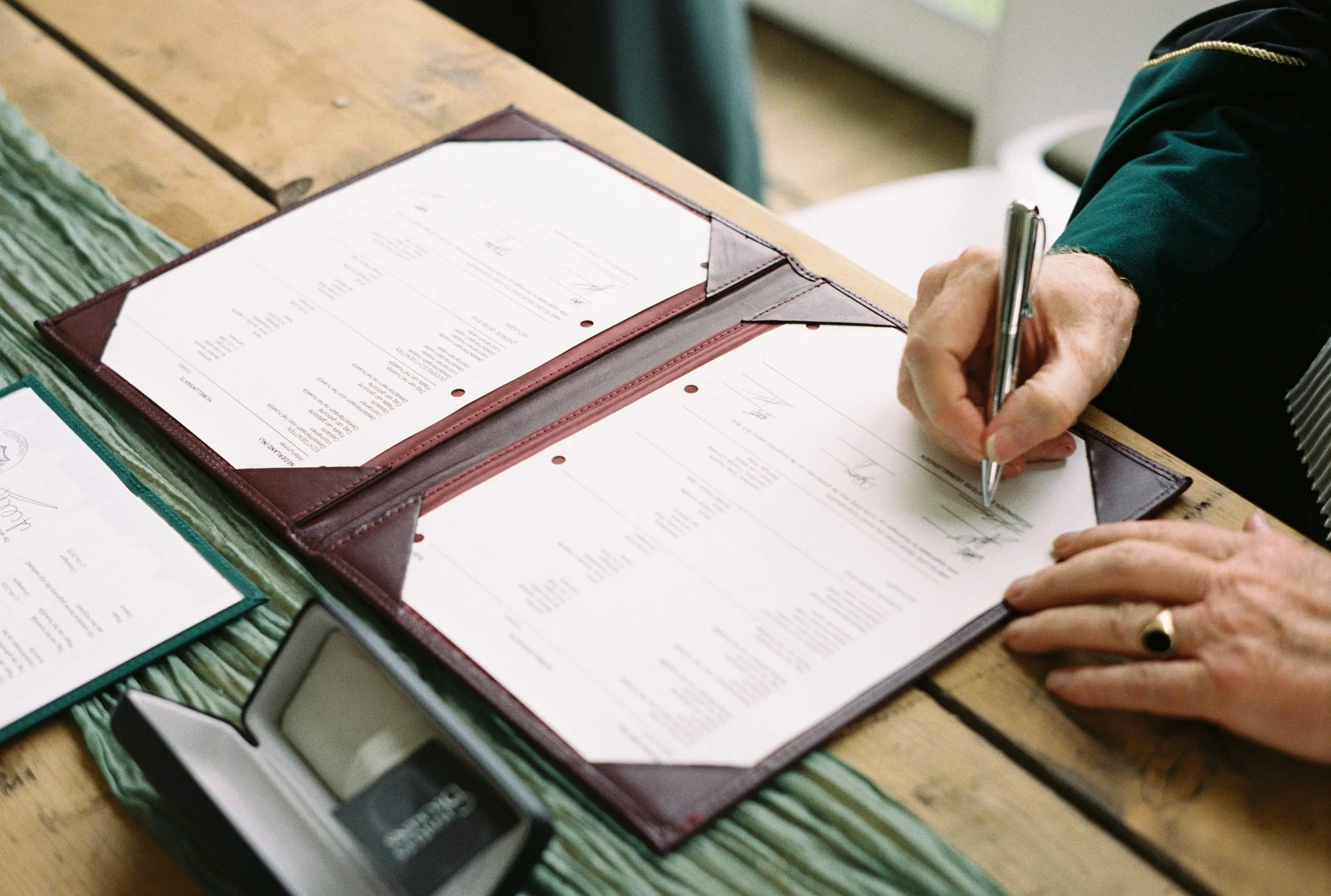 Person signing papers on a wooden table with documents in a leather folder.