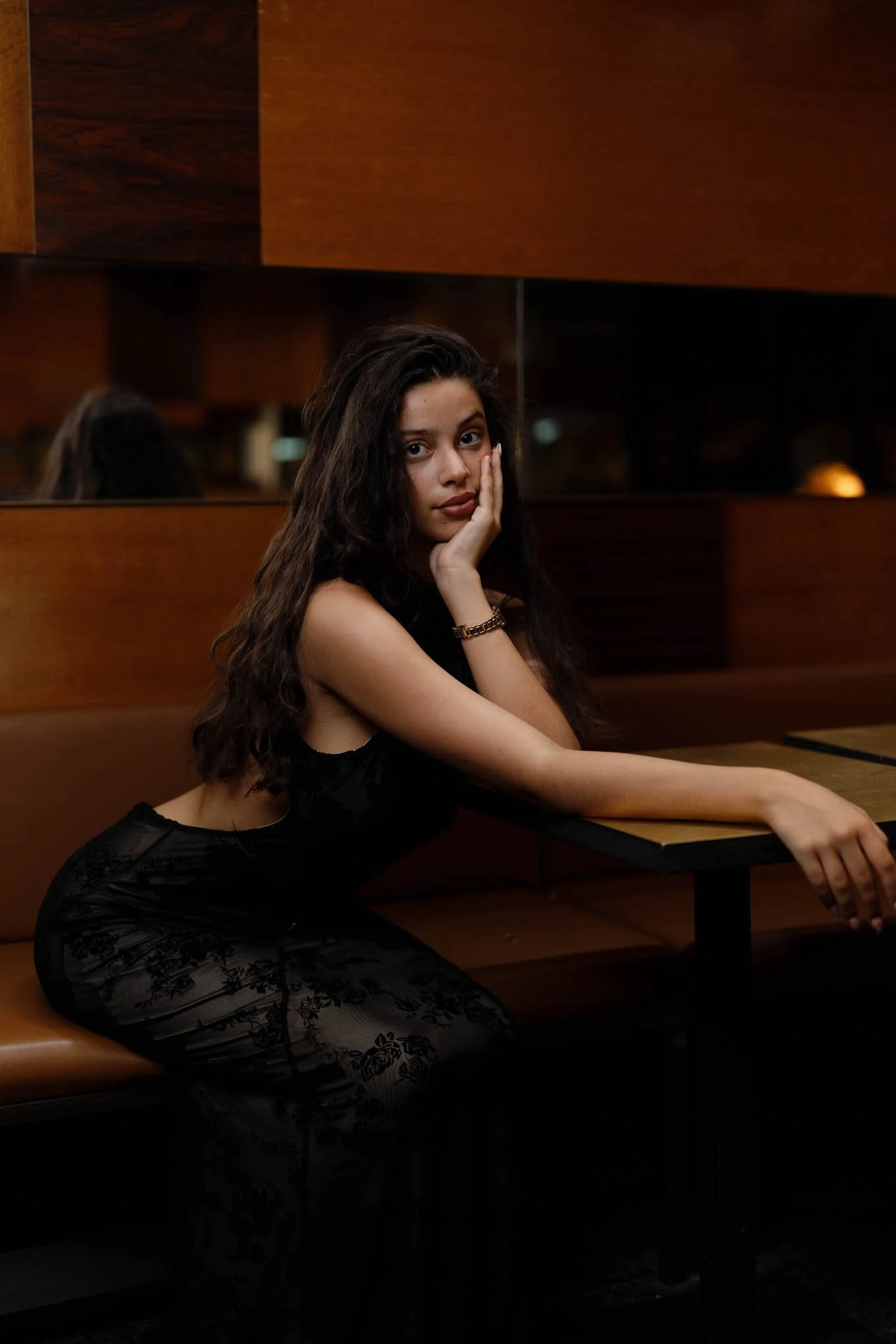 A young woman with long, curly brown hair, wearing a black lace dress, sitting in a dimly lit room with wooden decor, resting her face on her hand, looking into the camera.