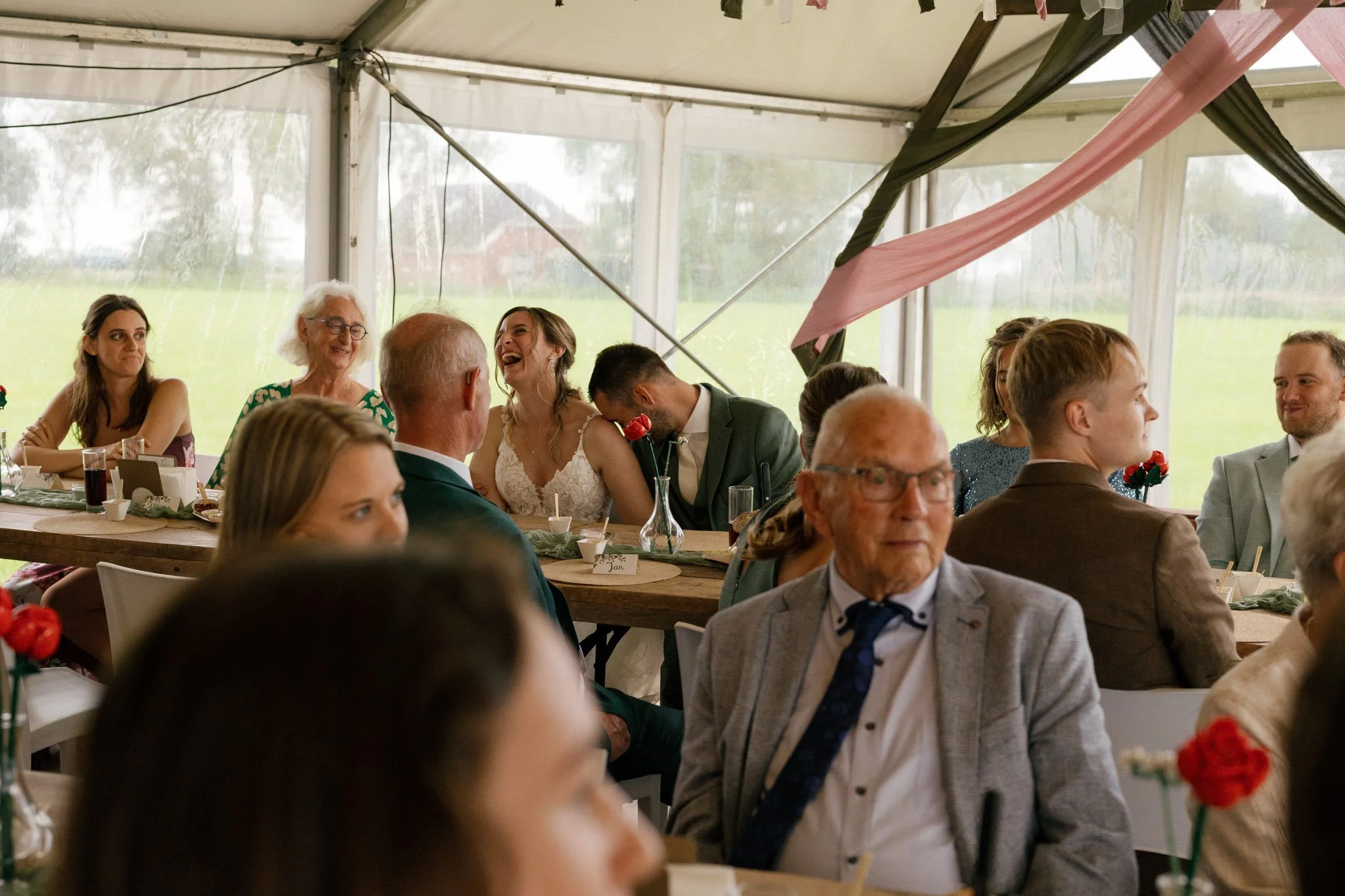 Guests at a wedding reception, some laughing and smiling, seated at tables under a tent with transparent walls, while a bride and groom share a tender moment.