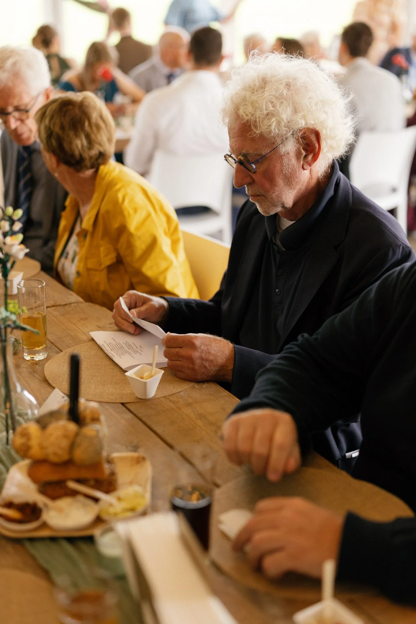 People sitting at a long table with food and drinks during a social gathering or party, in a well-lit room with a warm atmosphere.