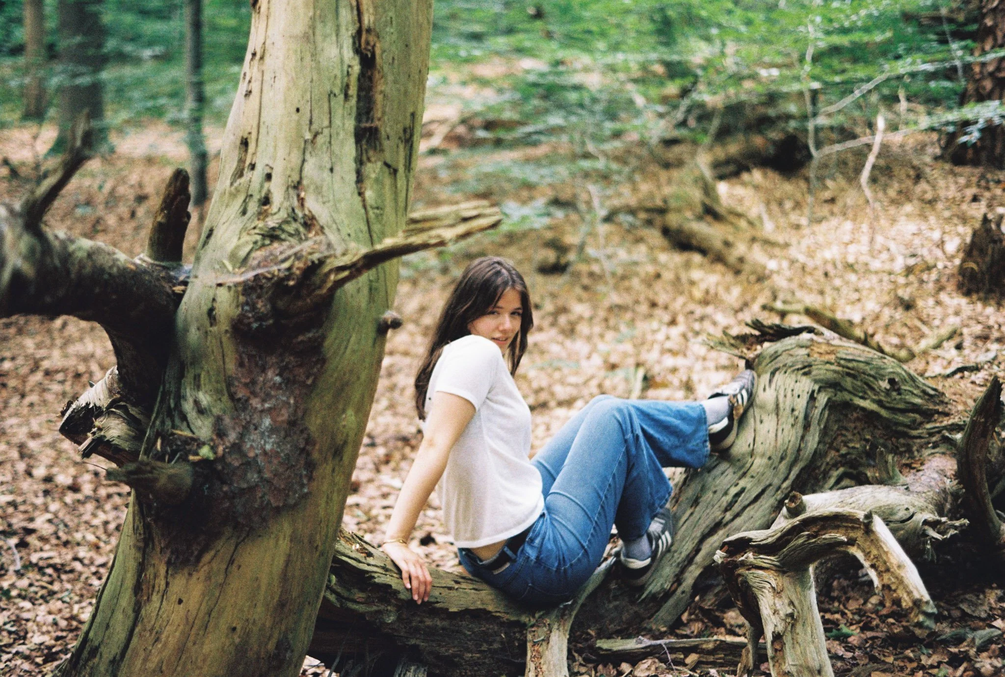 A woman with long brown hair sitting in a fallen tree in a forest, smiling at the camera with a natural background of trees and leaves.