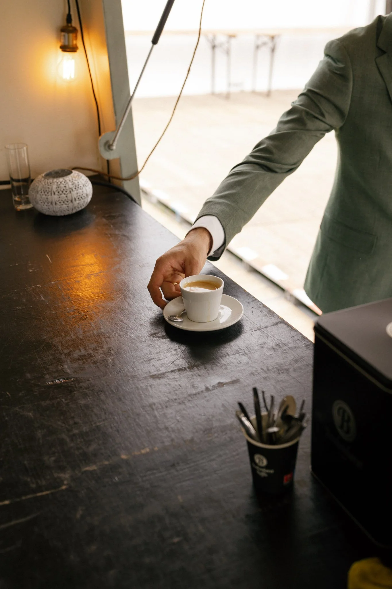 A person in a gray suit is serving coffee from a white cup on a saucer at a dark wooden table. There is a small white decorative lamp and a glass of water on the table, with a container of black utensils nearby. The background shows an outdoor area with a bright space and ornate columns.