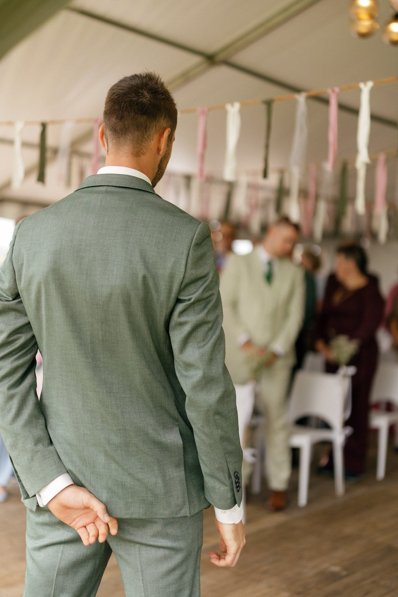 A man dressed in a gray suit standing with his hands behind his back while attending a formal event, with blurred people in formal attire in the background.