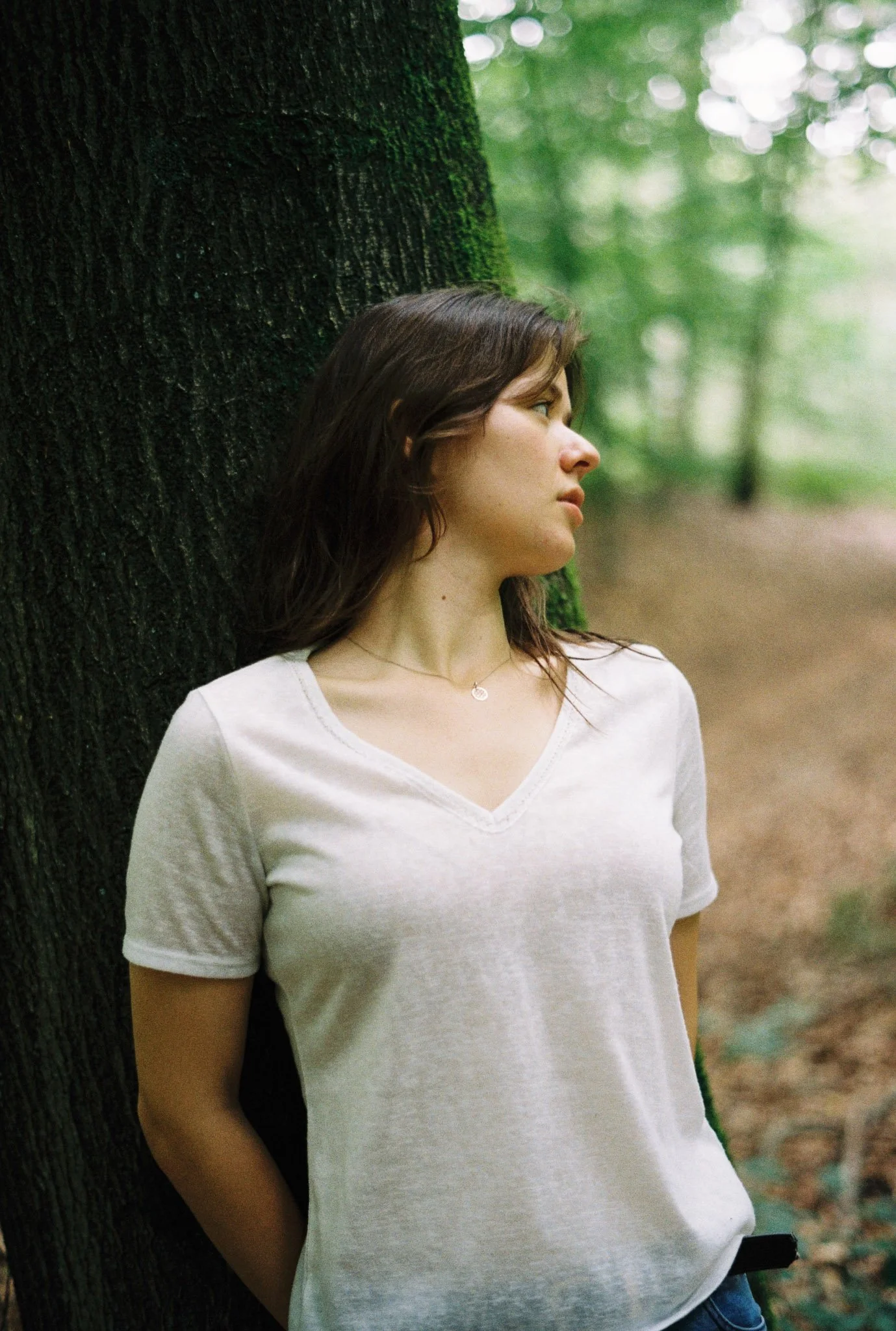 A young woman with brown hair wearing a white t-shirt leaning against a tree in a green forest.