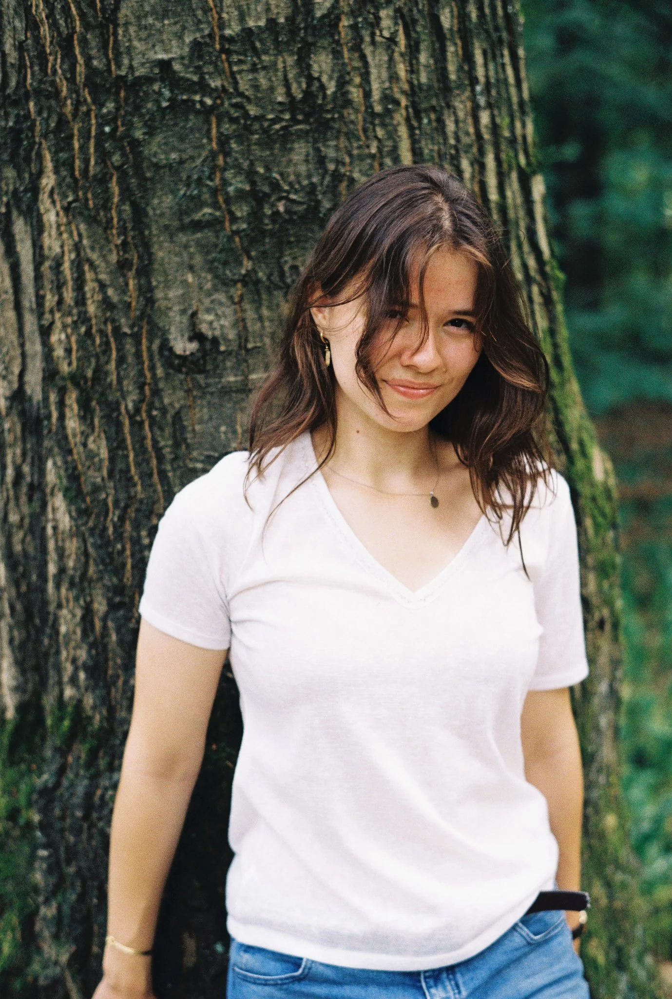 A young woman with wavy brown hair, wearing a white t-shirt, gold earrings, a necklace, and a bracelet, standing in front of a large tree trunk in a forest setting.
