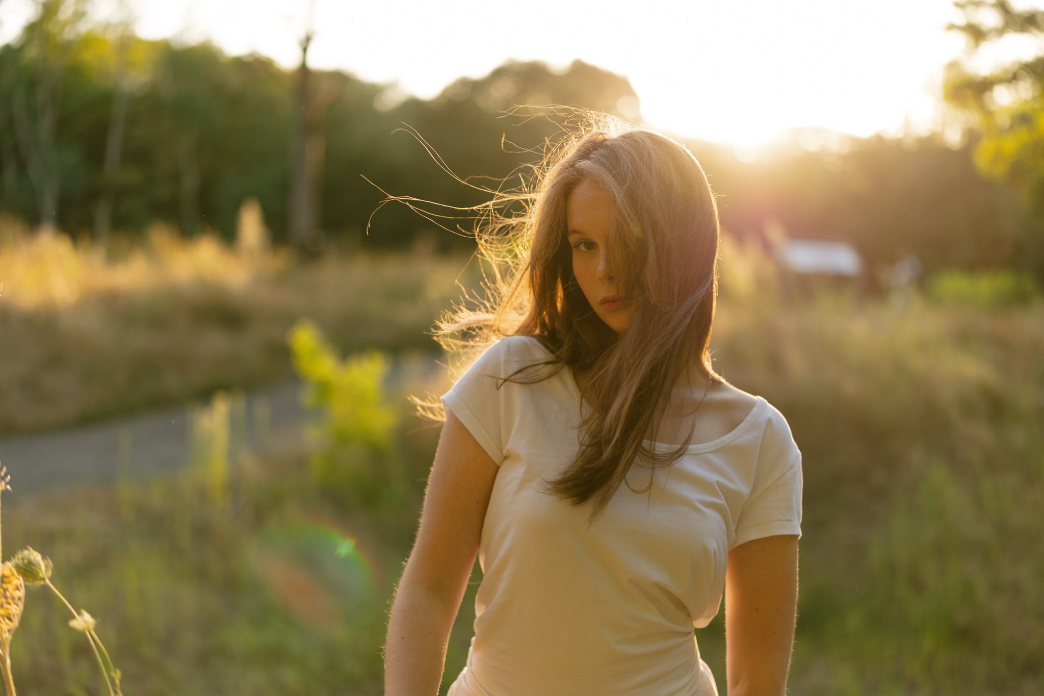 A young woman with long hair standing outdoors during sunset, wearing a white t-shirt, with trees in the background.