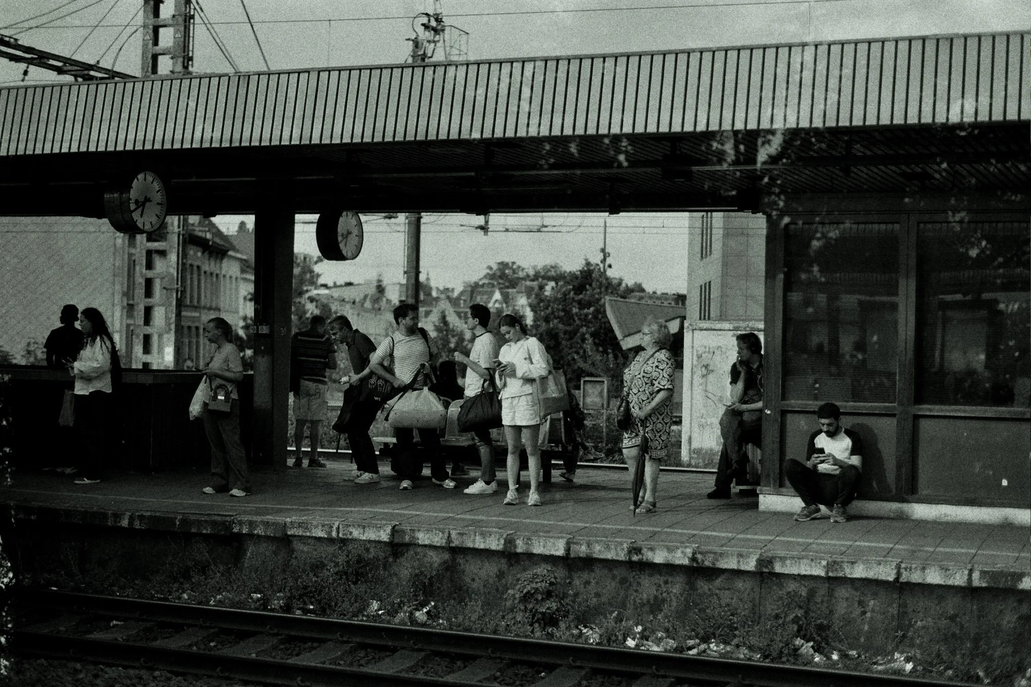 People waiting at a train station platform with luggage, some chatting and some sitting, under a roof with clocks showing the time. Train tracks are visible in the foreground.