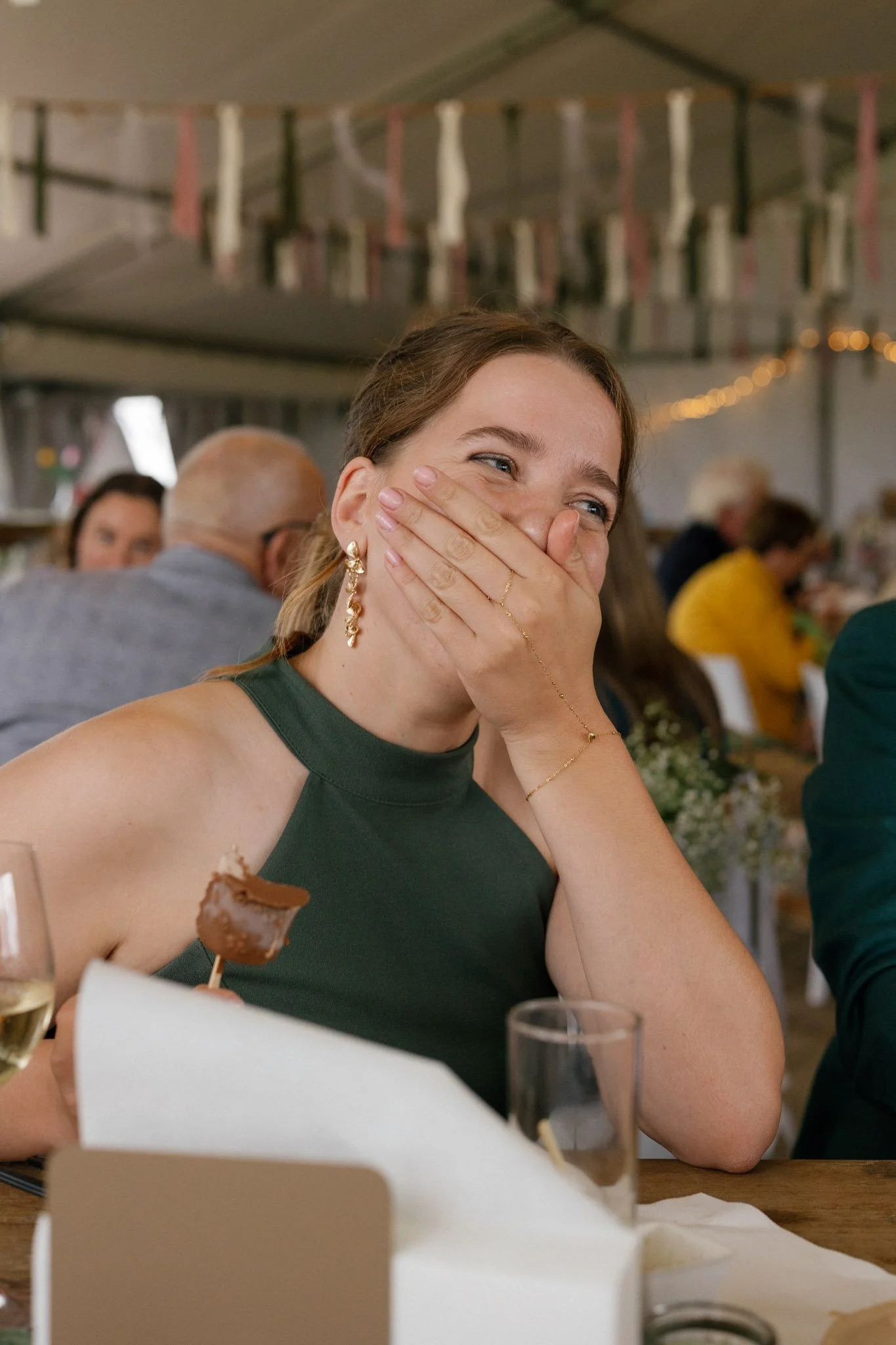 A woman with brown hair, wearing a green sleeveless dress and gold jewelry, is smiling and covering her mouth with her hand during a celebration or gathering inside a decorated tent.