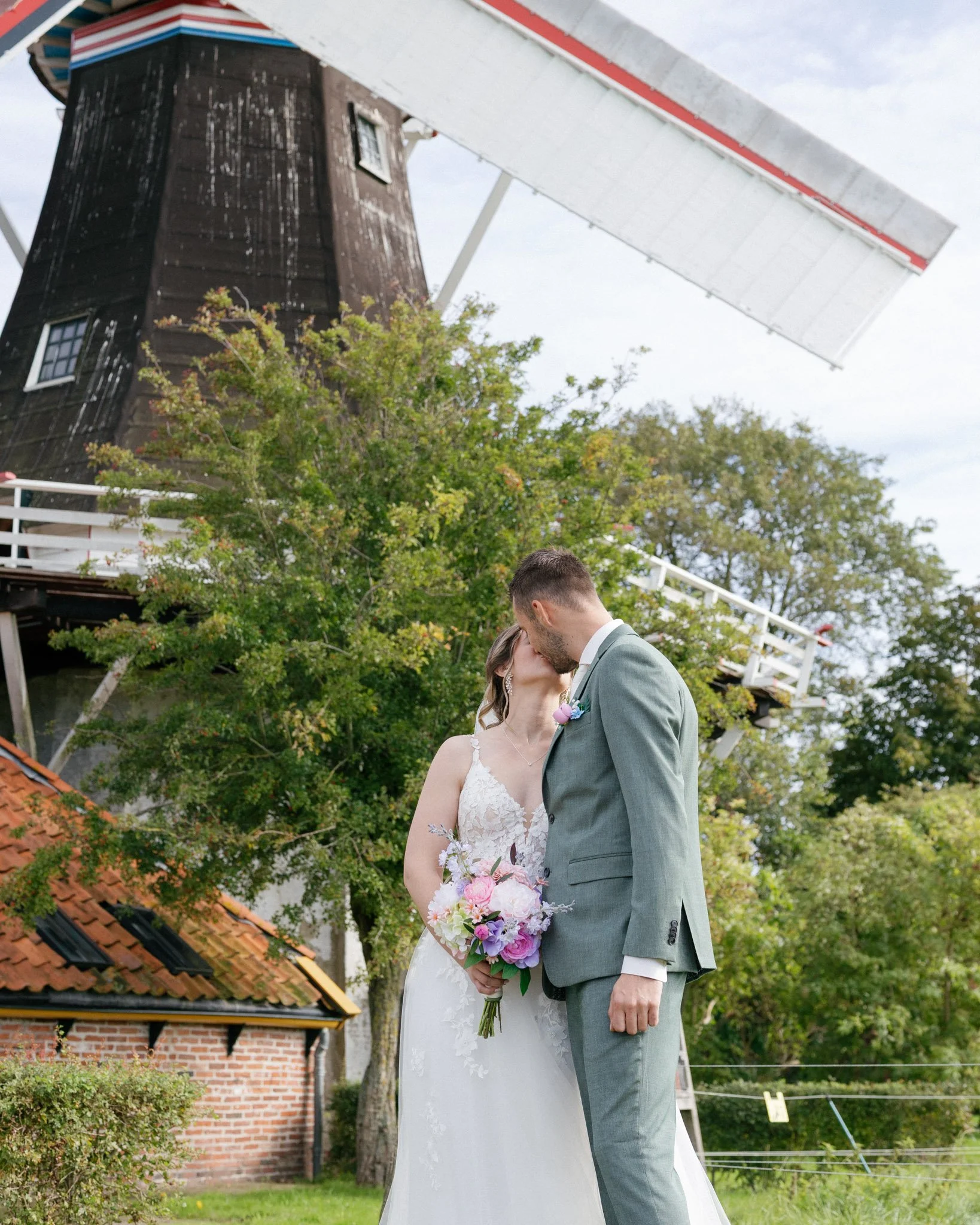 A bride and groom share a kiss outdoors during a wedding ceremony, with a windmill and lush green trees in the background.