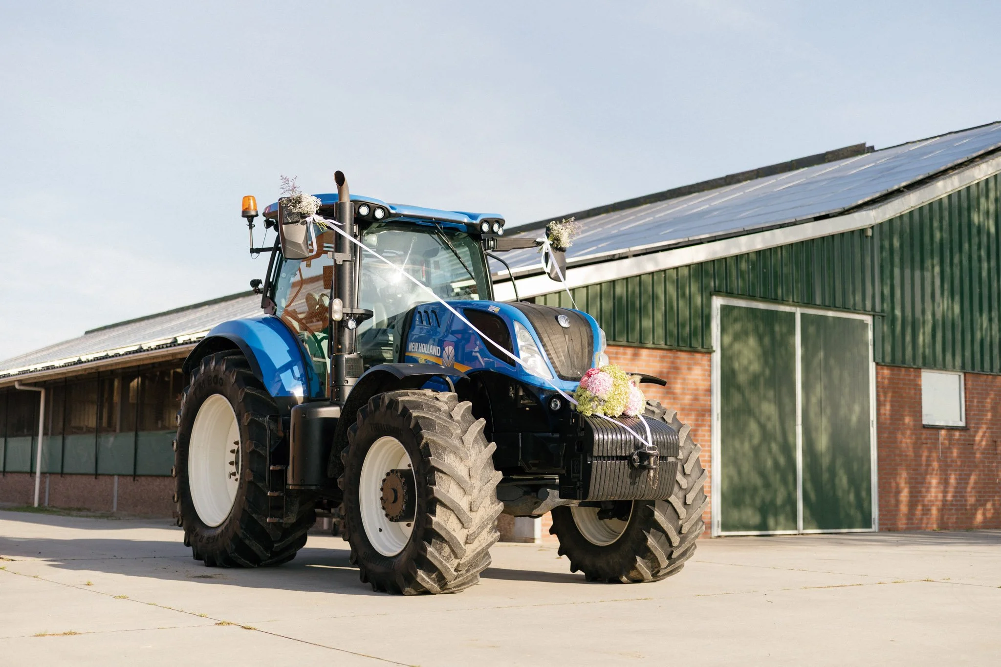 A blue tractor decorated with flowers and ribbons, parked outdoors on concrete, with a green building and a clear sky in the background.