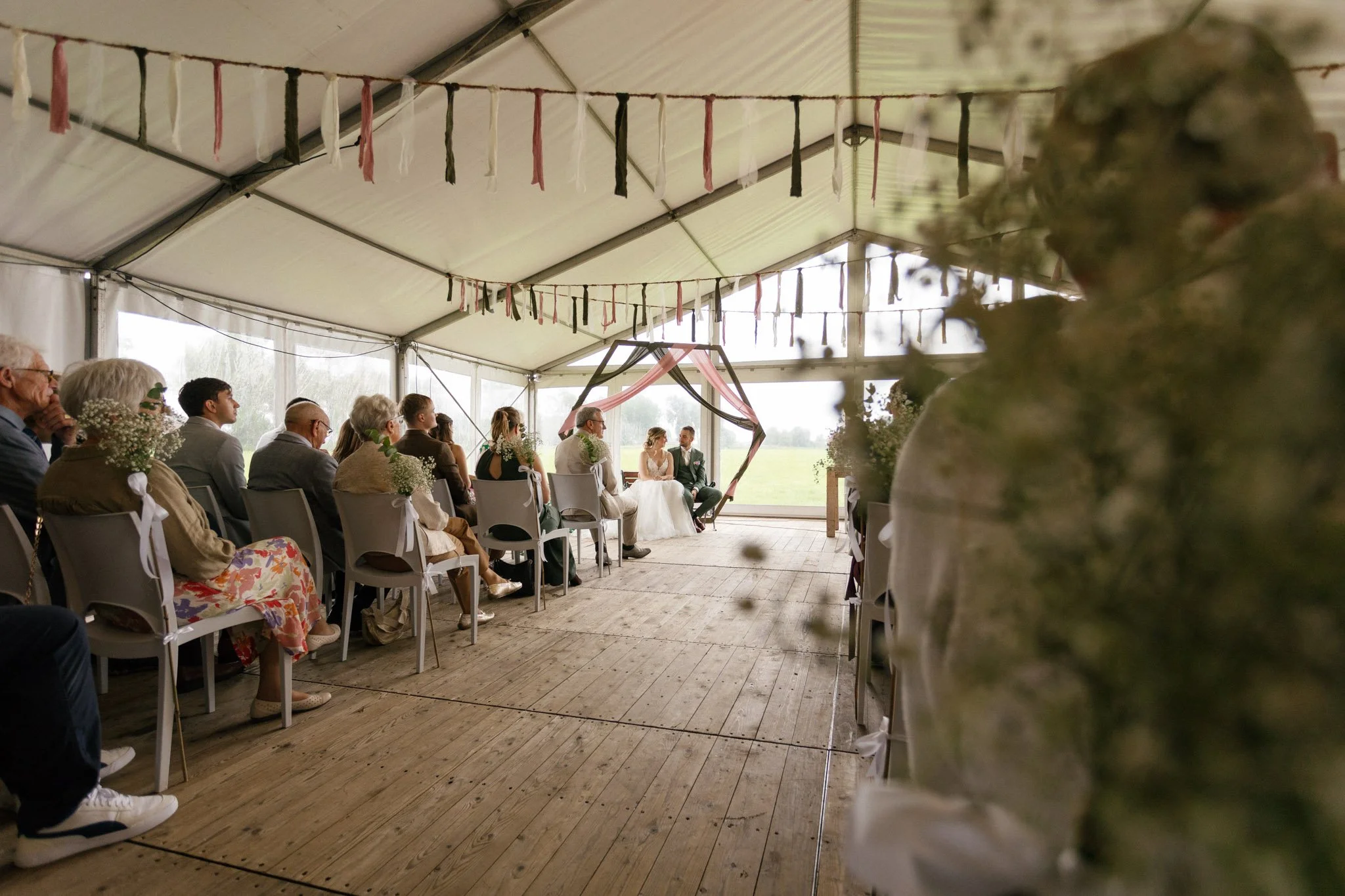 Wedding ceremony inside a large tent with guests seated, bride and groom seated at the front, decorative fabric and string decorations hanging from the ceiling, and flowers in the foreground.