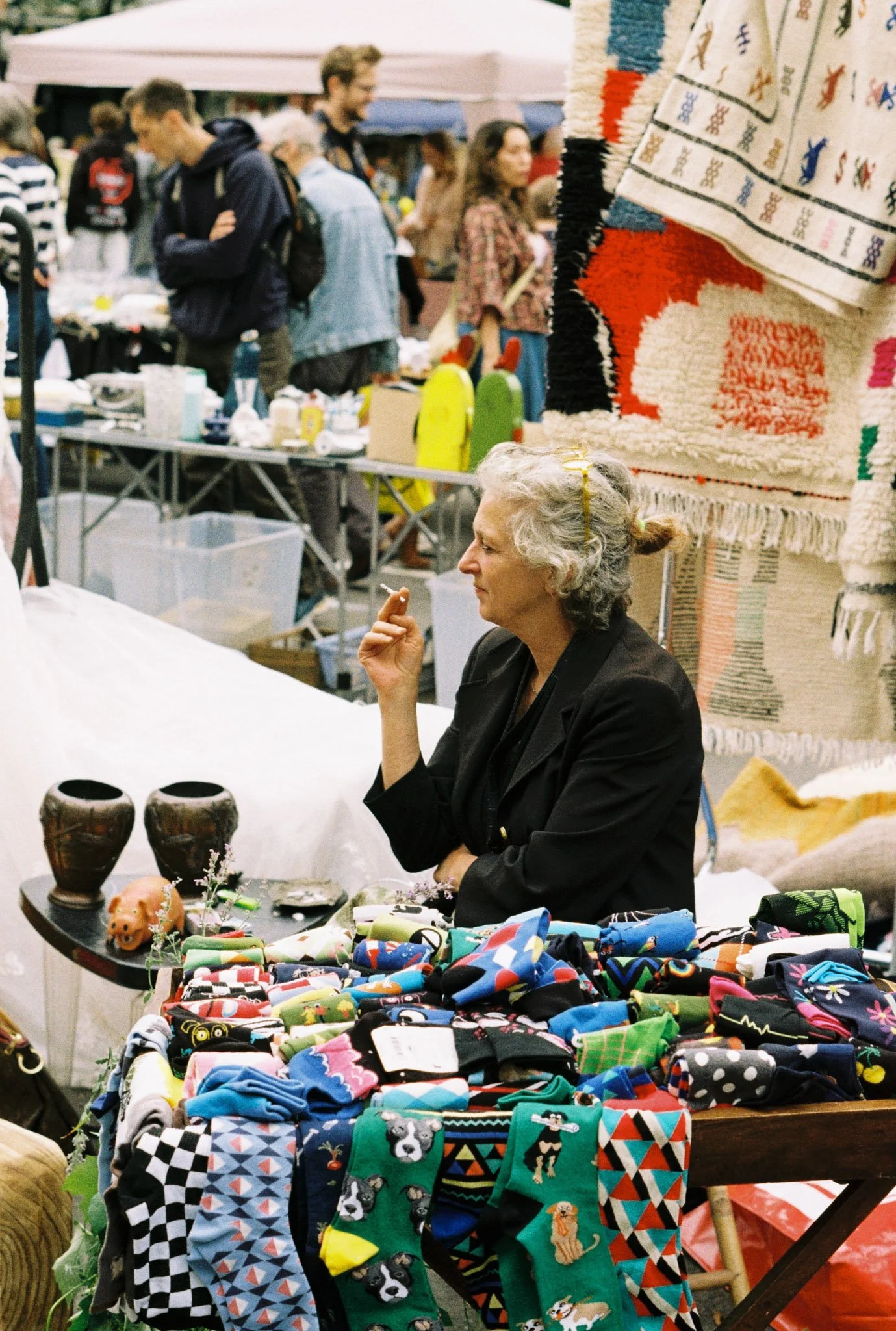 A woman sitting at a table with colorful socks featuring various patterns and animals at an outdoor market, with other shoppers and stalls visible in the background.