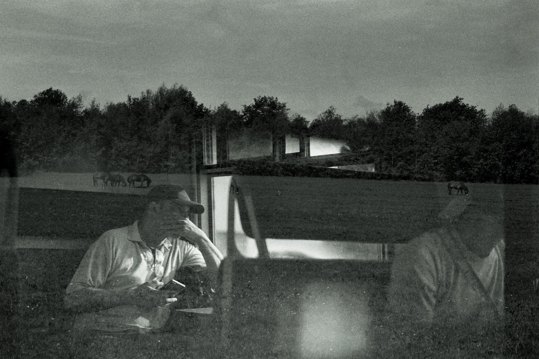 Black and white photo of a man sitting in a vehicle, looking at a phone, with trees, a bridge, and stars in the night sky visible through the window.