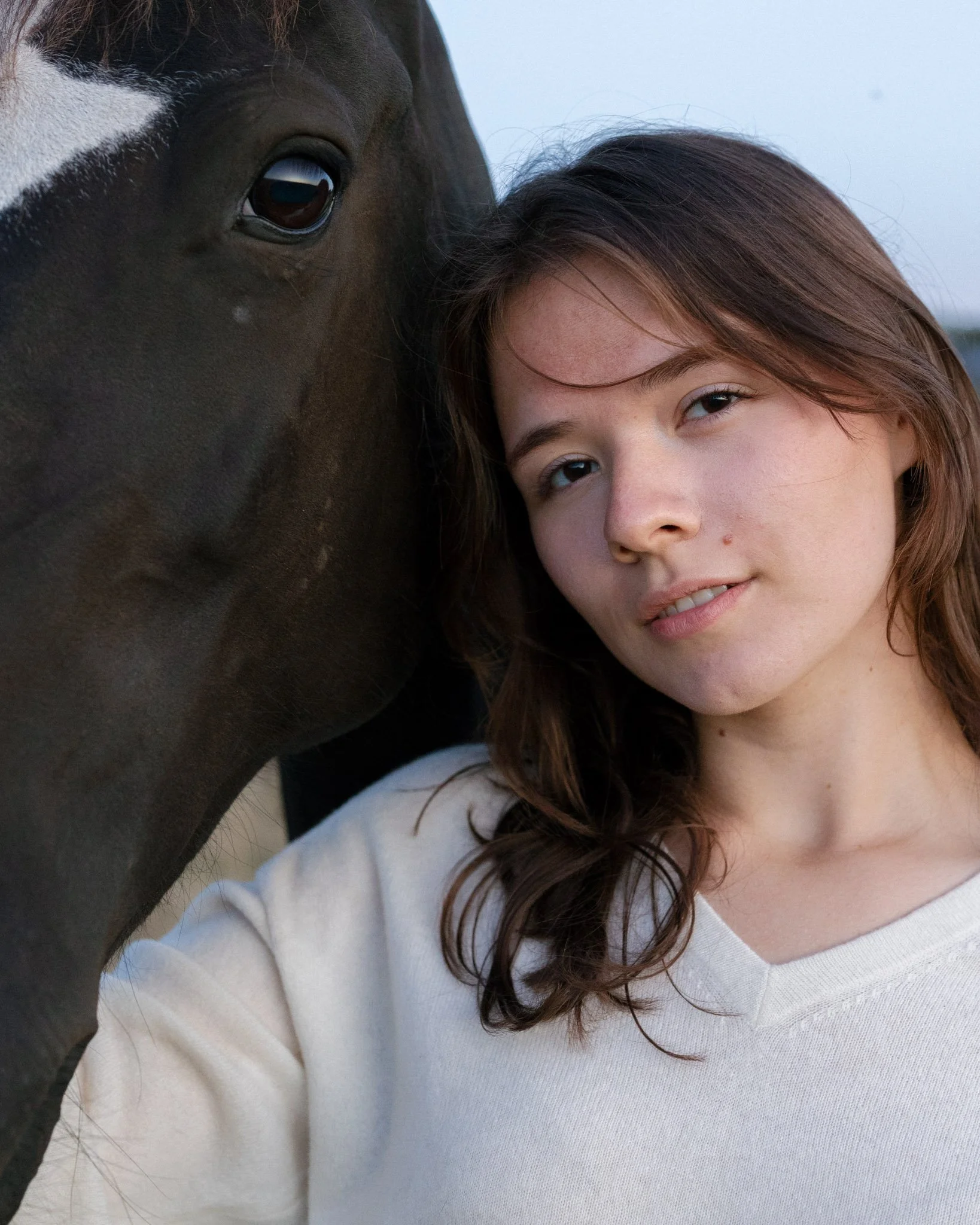 A close-up of a person with long brown hair and a white top, with their face partially resting against a horse's face. The horse has a dark brown coat with a white stripe on its face, and is looking at the camera.