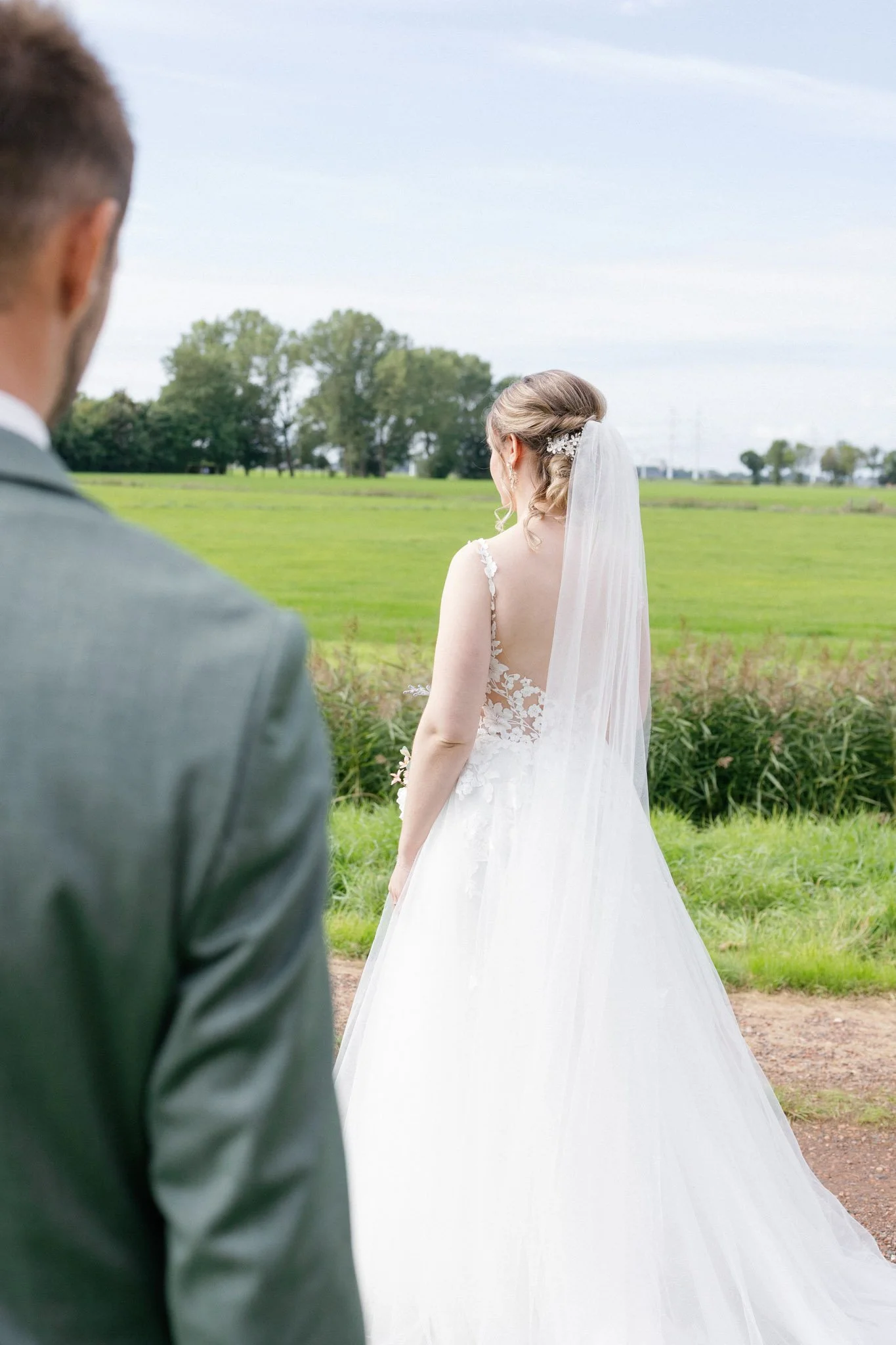 A bride in a white wedding dress and veil stands outdoors in a field with greenery and trees, with a groom in a gray suit visible in the foreground.