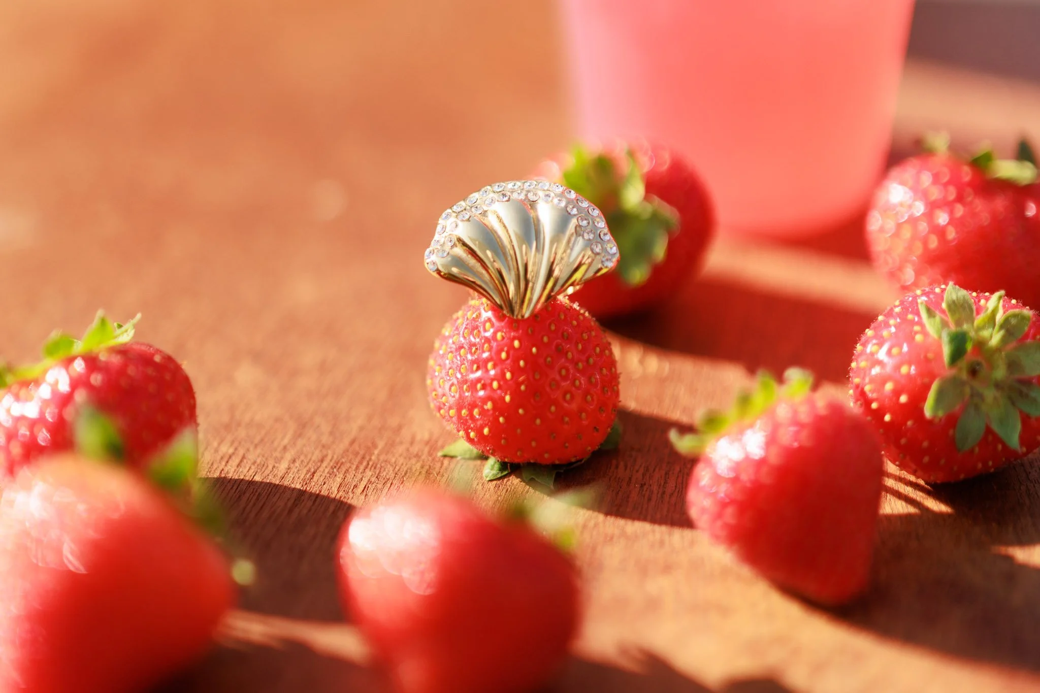Close-up of a silver seashell-shaped ring with rhinestones on a red strawberry surrounded by other strawberries and a pink cup in the background.