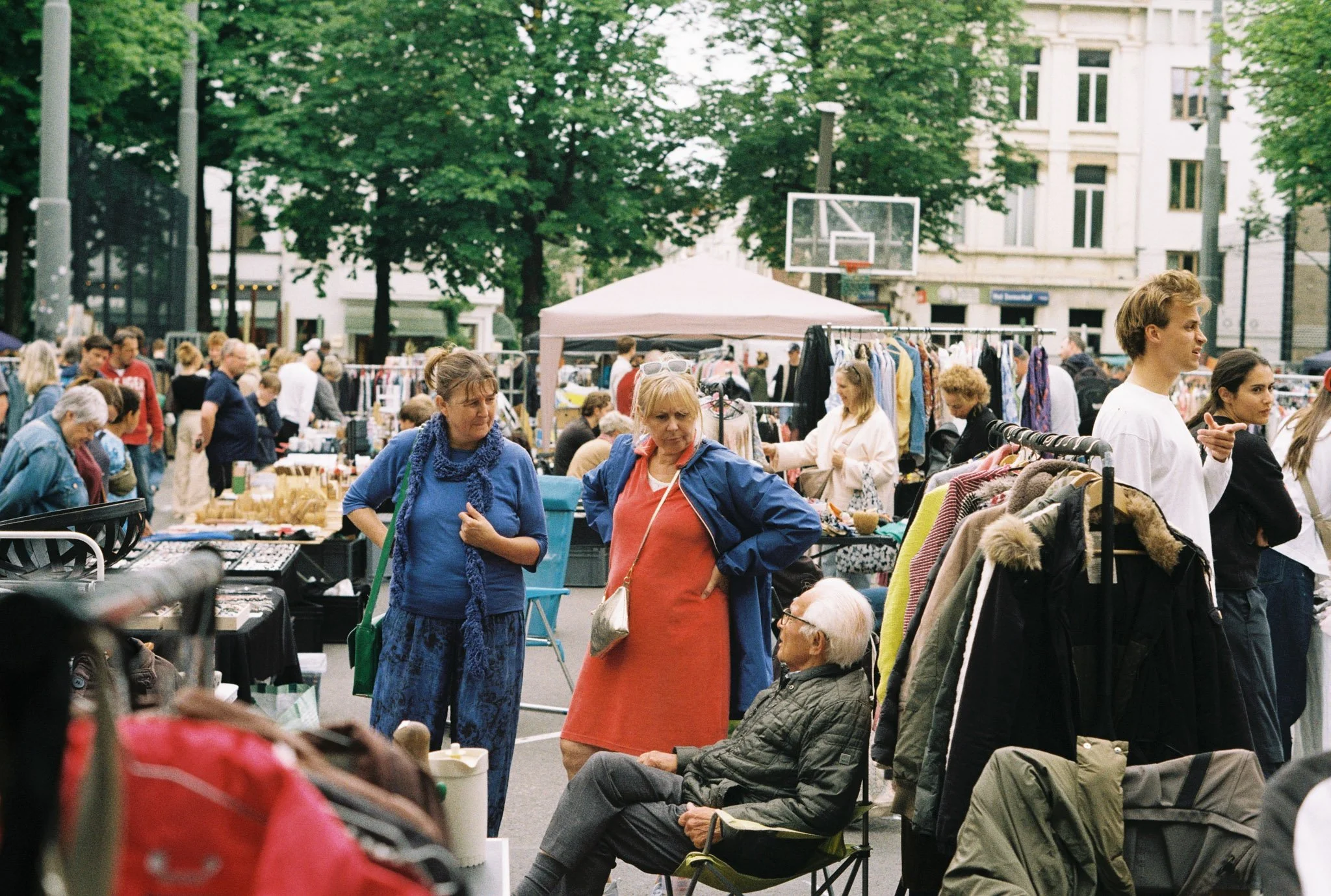 People browsing and shopping at an outdoor street market with racks of clothes and tables of items, under trees and buildings.