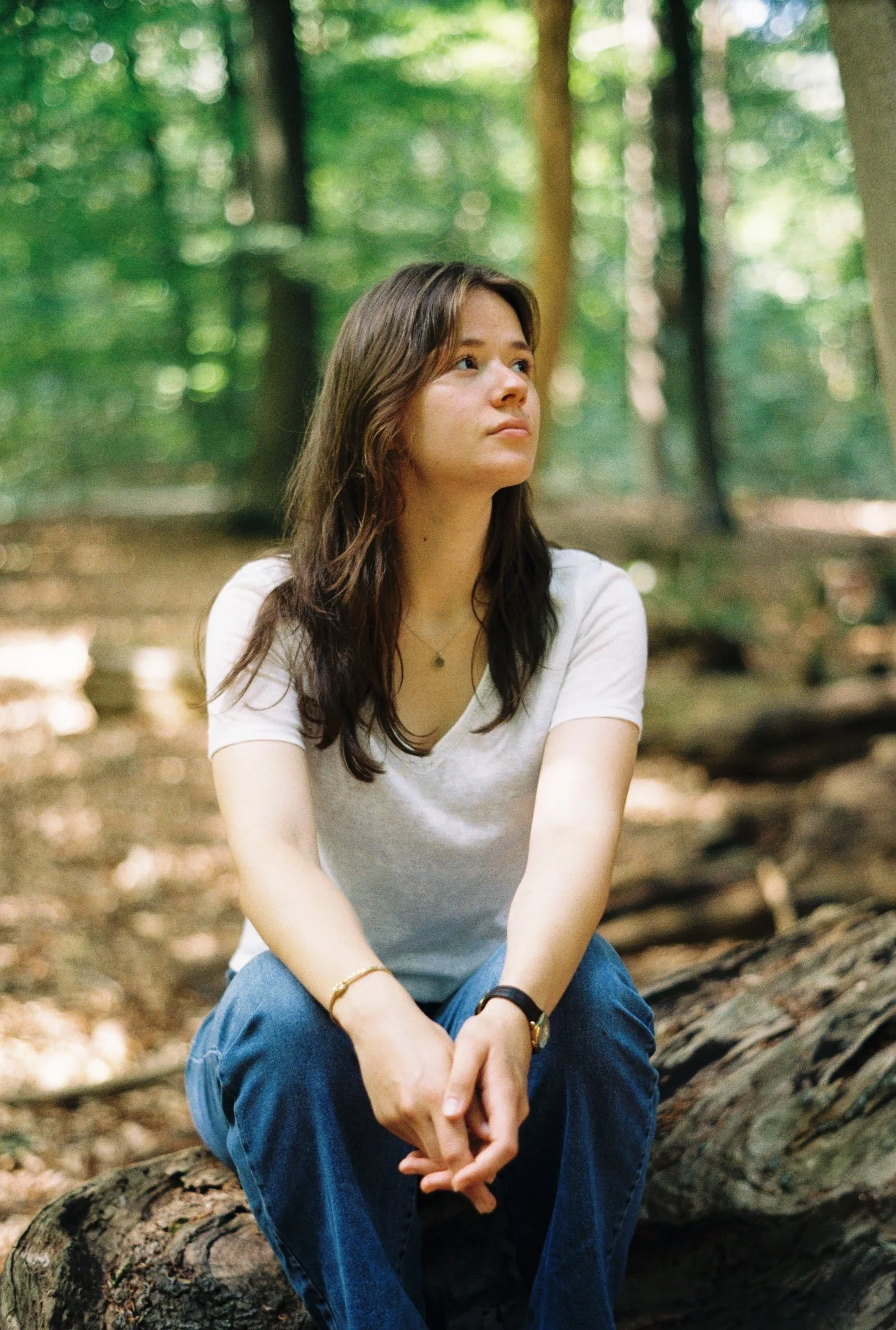 A young woman with brown hair sitting on a fallen log in a forested area with green trees in the background.